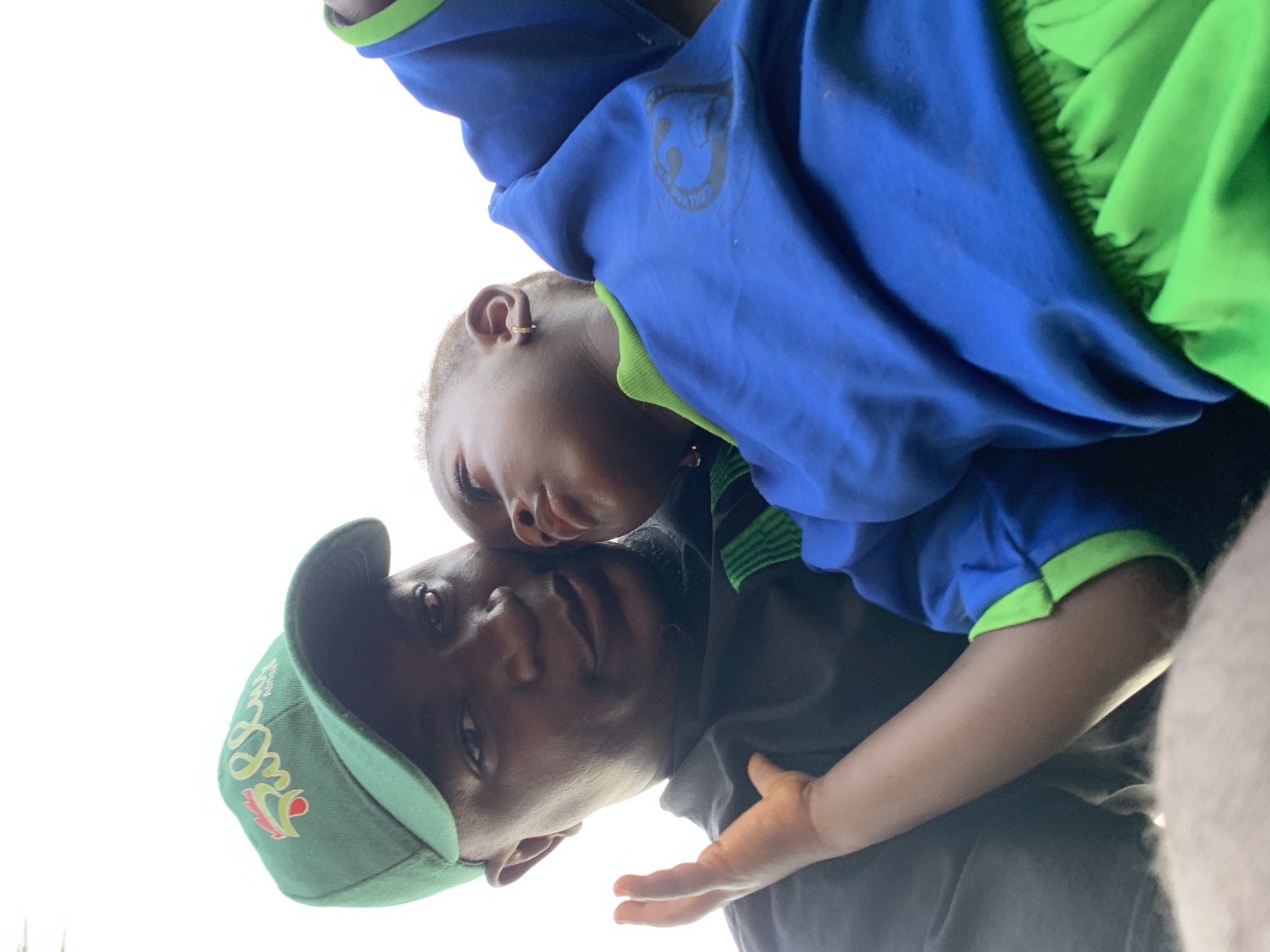 A photo taken from a low angle shows a man and a child in what appears to be Akwanga, Nigeria, based on the cap the man is wearing. The man wears a green cap with the word "Akwanga" embroidered on it in yellow, red, and green. He also wears a dark shirt. The child, who appears to be resting their head on the man's head, is wearing a blue and green shirt or sweatshirt with a round emblem on the chest. The sky is visible in the background, suggesting an outdoor setting. The photo appears candid, possibly taken with a phone or similar device.