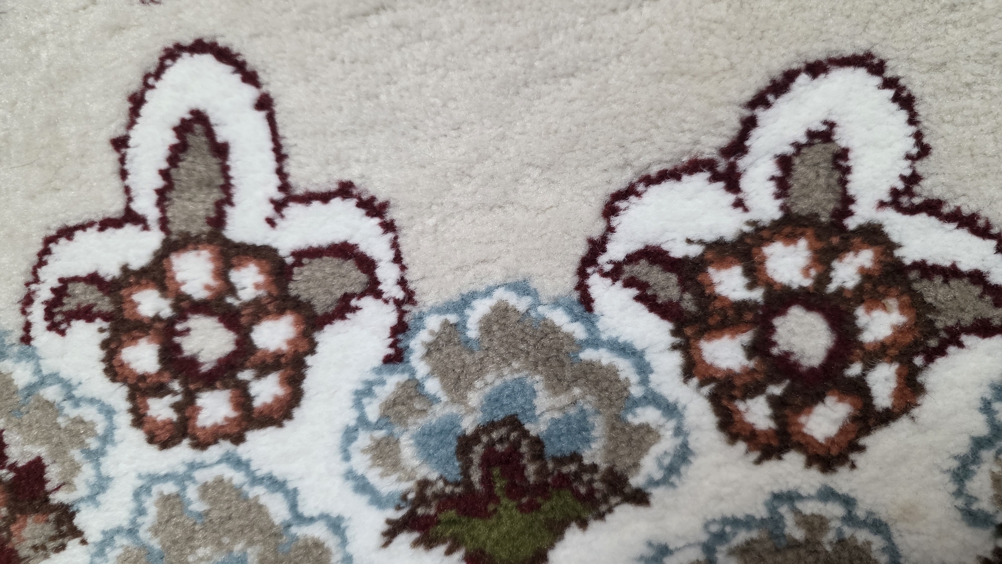 A close-up, overhead shot shows a cream-colored carpet with a repeating floral pattern. The floral motifs are composed of intricate designs in shades of burgundy, white, and muted blue and green. The carpet appears to be soft and plush, with visible texture from the woven fibers. The lighting is even, highlighting the details of the pattern. The image is cropped tightly on the carpet, and there is no visible indication of its location in Chuy, Kyrgyzstan.