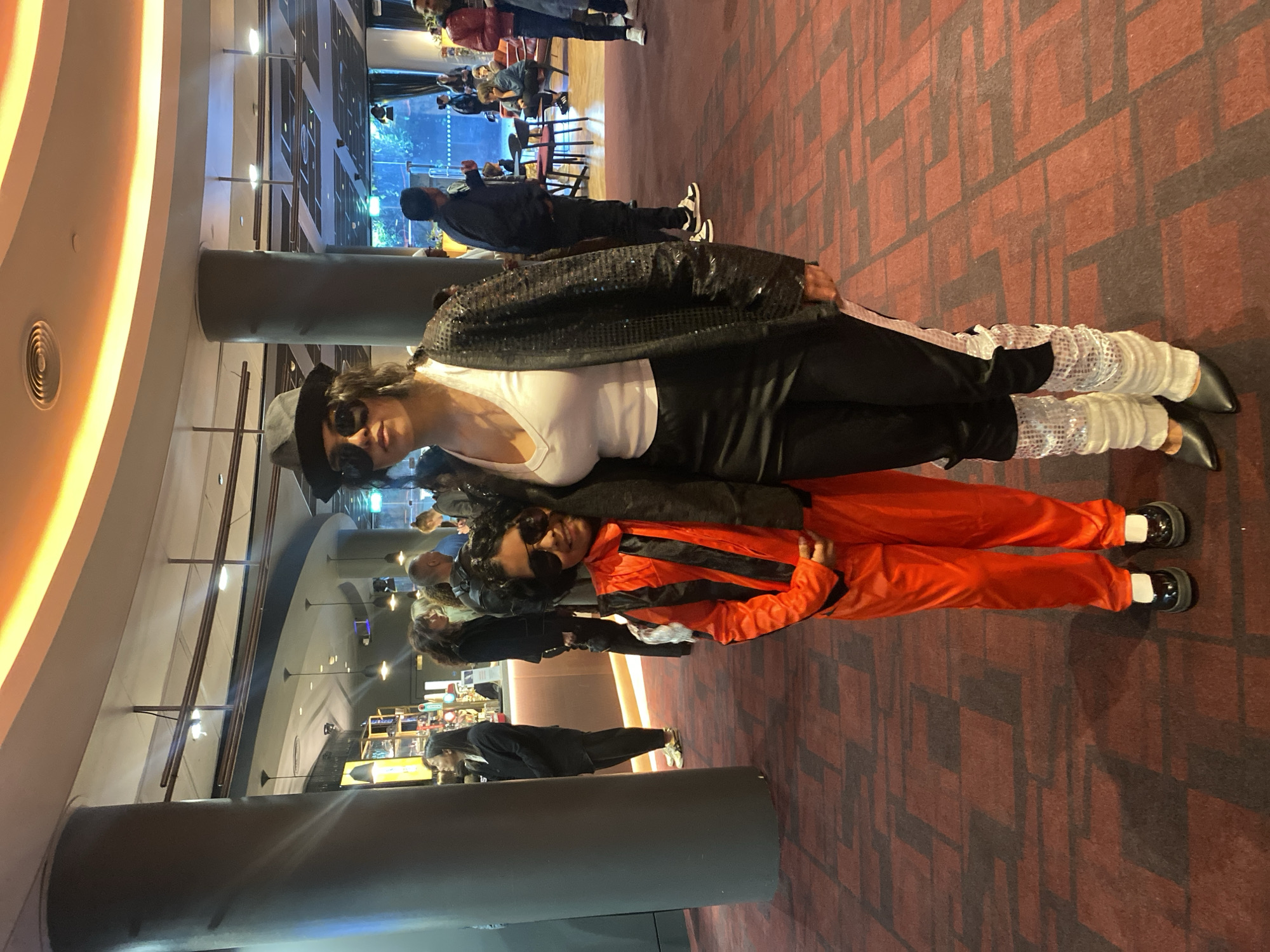 The image captures two individuals in elaborate costumes posing indoors, likely within a public entertainment venue in London, United Kingdom, during the evening.
In the foreground, a taller person on the left is dressed in a Michael Jackson-inspired outfit. They wear a black fedora, dark sunglasses, a white V-neck top, a glistening black sequined jacket, black trousers, and distinctive white sparkly leg warmers over black pointed shoes. Their dark, curly hair peeks out from under the hat.
Adjacent to them, a shorter individual, likely a child, smiles while wearing an orange jumpsuit with prominent black stripes, possibly reminiscent of a racing suit or a specific character's attire. This person also wears dark sunglasses and black shoes with white socks.
The setting is characterized by a spacious interior featuring a red and dark geometric-patterned carpet and large dark pillars. In the background, numerous casually dressed people are visible, some seated at tables and others standing, indicating a social or waiting area. Overhead, warm artificial lights curve along the ceiling, and various downlights illuminate the space, suggesting a nighttime or indoor ambiance. The costumed individuals appear to be enjoying themselves, possibly attending a themed event or an entertainment outing. No visible text is discernible within the image.