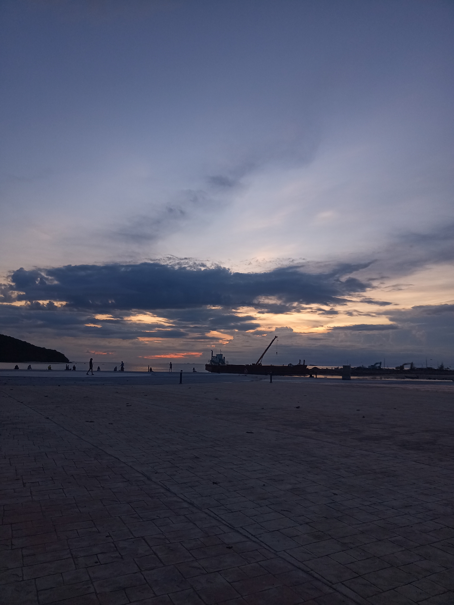 The image depicts a twilight scene at a coastal area, likely a promenade or pier, in Koh Phangan, Thailand. The sky is a gradient of deep blue to lighter shades of purple and grey, with streaks of orange and yellow from the setting sun visible through dark, textured clouds. A body of water stretches out to the horizon, reflecting the soft light. On the left side of the frame, a dark, hilly silhouette suggests land.
In the foreground, a paved area with a pattern of interlocking rectangular stones leads towards the water. Scattered across this area and closer to the water's edge are numerous silhouetted figures of people, some appearing to be walking or standing, while others are seated. Their forms are indistinct due to the low light.
Further into the scene, on the water, are several large vessels. One prominent object is a ship with a visible crane, appearing to be docked or anchored. In the distance, other structures that might be port facilities or more ships are faintly visible. The overall atmosphere is serene, with the fading light casting long shadows and creating a moody ambiance.
