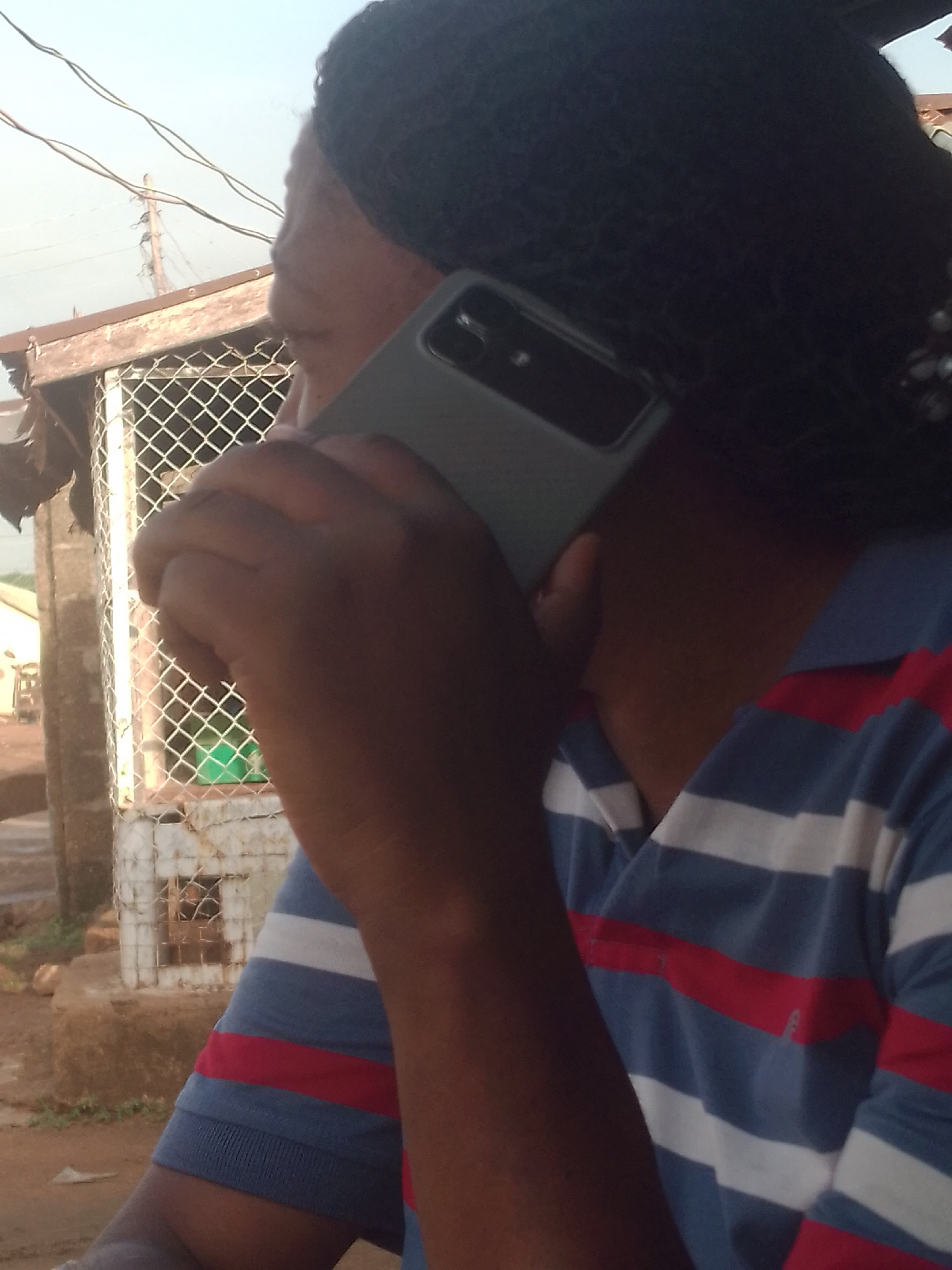 The image captures a close-up, angled view of a person engaged in a phone call outdoors. The individual, who has dark skin, is seen from the side, with the left side of their face mostly obscured by a gray smartphone held to their right ear with their right hand. Their head is covered in dark, possibly braided hair or a headwrap. They are wearing a short-sleeved polo shirt with horizontal stripes in blue, white, and red.
In the background, there is a metallic mesh fence or cage-like structure, possibly part of a building or enclosure, topped with wooden beams. Overhead, several utility wires are visible against a bright, clear sky, suggesting an outdoor, daytime setting. The ground appears to be unpaved, possibly dirt or rough concrete. The lighting indicates it is daytime, likely mid-morning or afternoon, with soft, natural light. No specific emotions are discernible from the visible portion of the person's face, and there is no visible text in the image. The overall setting appears to be an informal or semi-urban environment, possibly in a developing area, given the construction and utility elements.