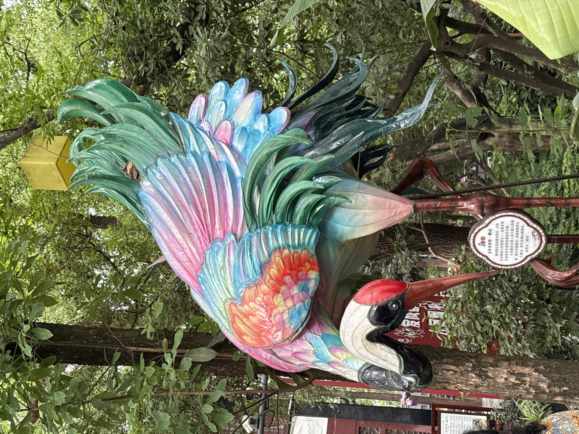 The image captures a vibrant, colorful crane sculpture set amongst lush greenery, likely within a park or garden. The crane's plumage is painted in a mix of blues, pinks, reds, and greens, giving it a whimsical appearance. A yellow cube or block is located near the tail of the crane. Next to the bird sculpture is a sign with information written in Chinese. This sign mentions the crane is called the "Heming" teahouse, located in the Qingyang District, China. The foliage surrounding the sculpture suggests a natural, outdoor setting. The camera angle is slightly tilted, and there are people and possible other bird sculptures located on the ground, lower portion of the image.