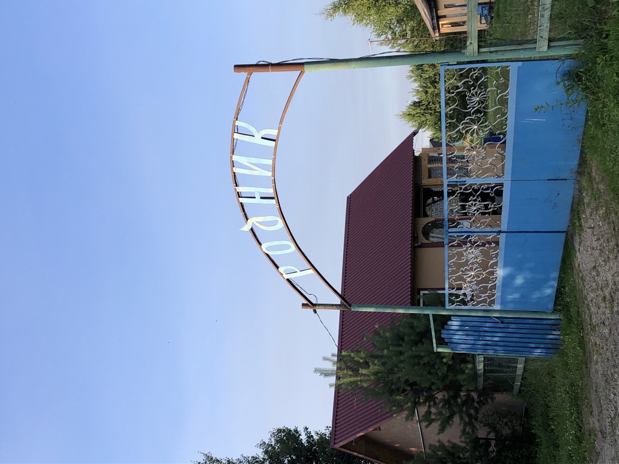 The image shows an arched metal sign with Cyrillic lettering that reads "РОДНИК". The sign is supported by a rusty metal post on the right and another post partially obscured by a building on the left. Below the sign, there is a blue metal gate with decorative scrollwork on the upper half. To the left of the gate is a small blue wooden fence. In the background, a house with a reddish-brown roof is visible, surrounded by greenery. The scene appears to be in a rural or semi-rural setting, possibly a village or dacha community. The sky is clear and blue. Based on the sign's text, which translates to "Spring", and the overall appearance, this location is likely in Makeikha, Russia, possibly marking the entrance to a spring or a property named after one. There are no people visible in the image.