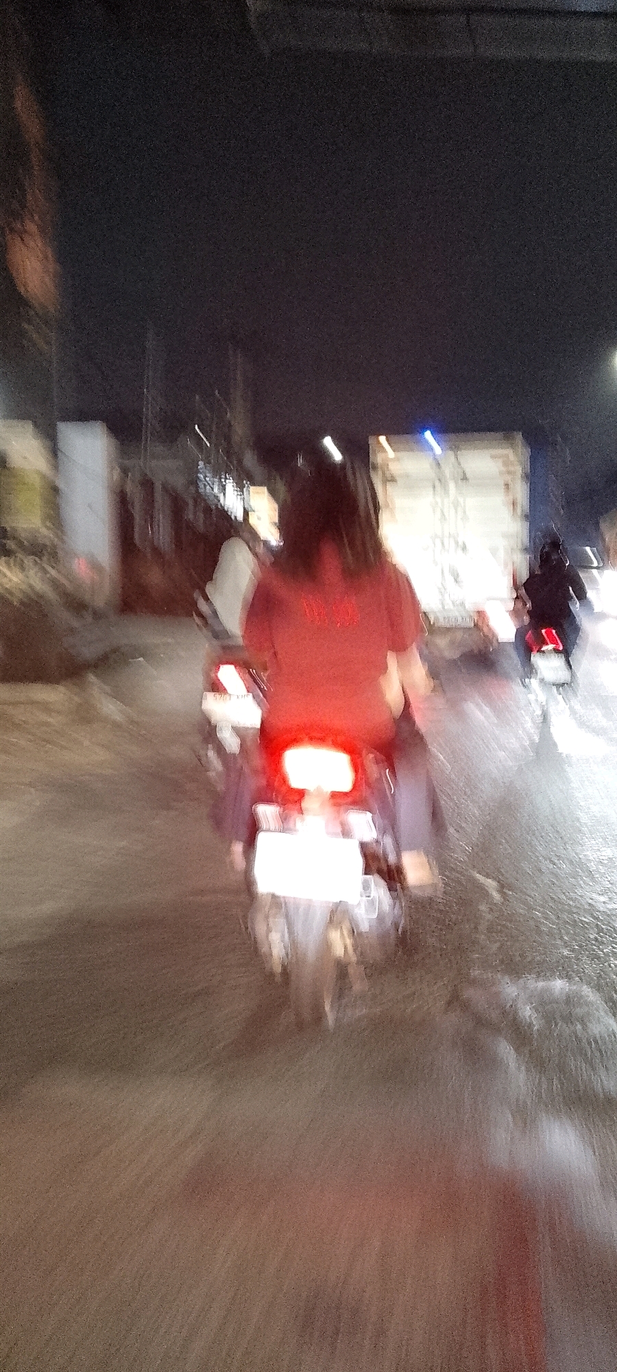 The image depicts a nighttime street scene in Bekasi, Indonesia, with motion blur indicating movement. In the foreground, a person wearing a red shirt is riding a motorcycle, with their face obscured. The motorcycle's red taillight is brightly illuminated, and a white license plate is visible. To the right of the foreground motorcycle, another motorcycle with a rider in dark clothing is also moving, its taillight illuminated red. In the background, a large white truck is visible with a blue light on its roof. The road surface appears wet and reflective, suggesting recent rain. Buildings and other urban elements are blurred in the background.