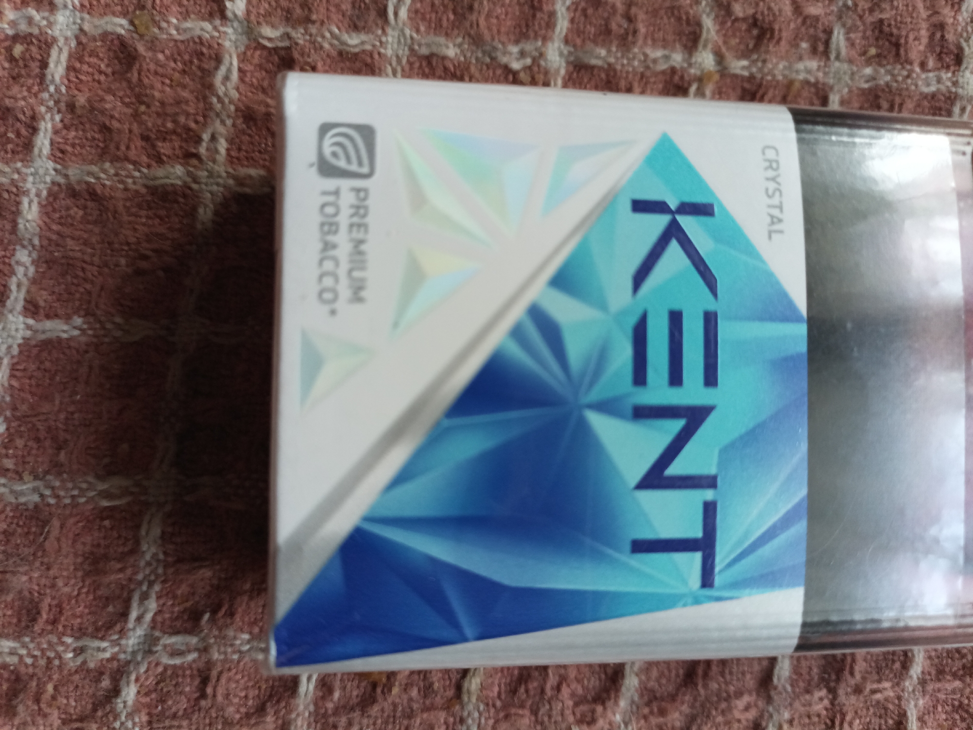 An overhead shot shows a closed pack of Kent cigarettes with a blue, geometric design and the word "KENT" in large, blue letters. To the left of the large letters, the words "PREMIUM TOBACCO" are printed vertically. To the right of the large letters, the word "CRYSTAL" is printed vertically. The pack is lying on a textured, reddish-brown surface.