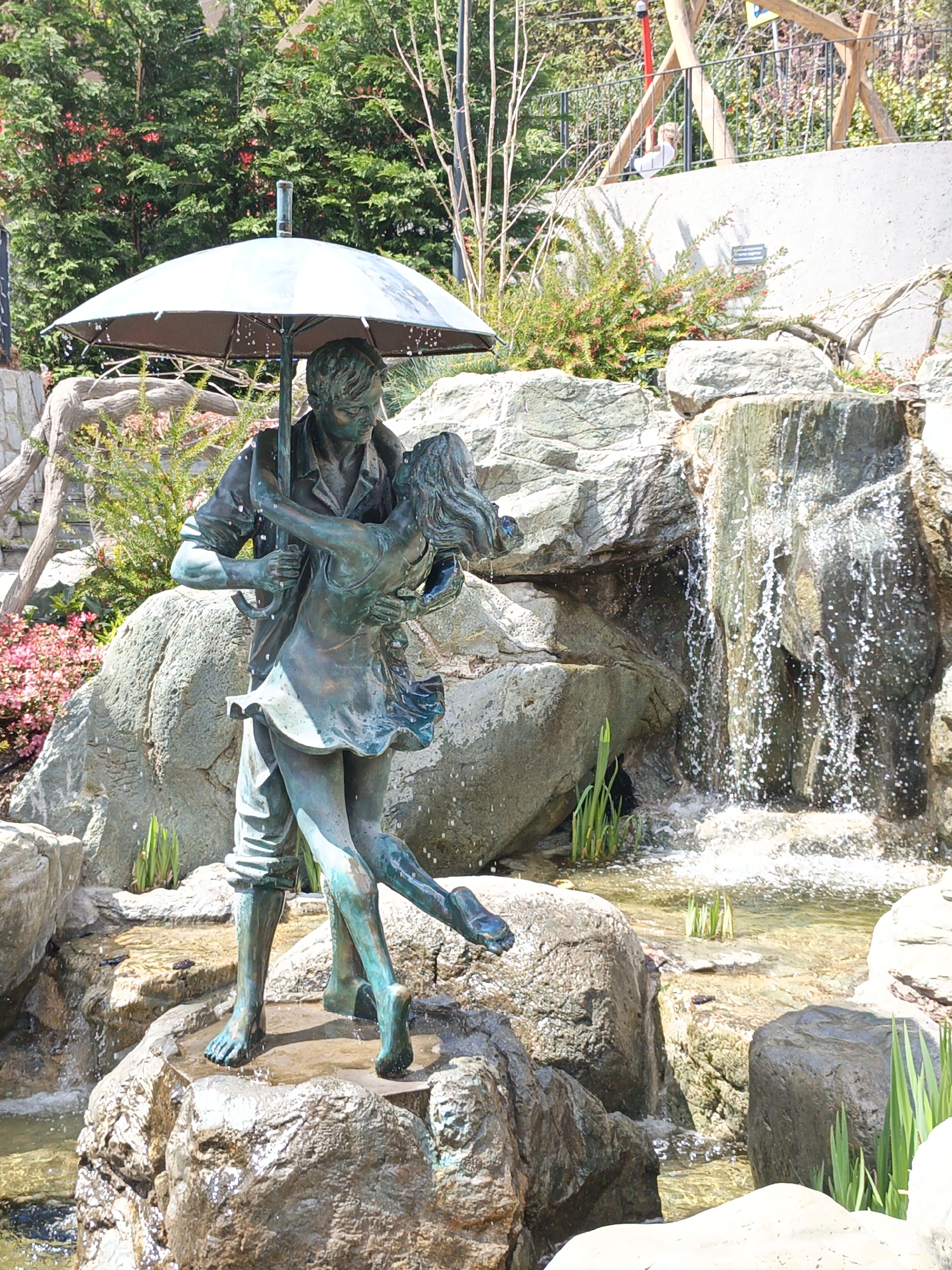The image is a photograph of a bronze statue depicting a couple dancing under an umbrella. The statue is located in a park or garden setting, likely near a body of water, as evidenced by the presence of rocks, water, and what appears to be a small waterfall in the background. The background also includes trees, plants, and a structure resembling a fence or barrier with people beyond. The statue appears to be the focal point of a park feature or water feature. The image was taken during daylight. The exact location in Верхореченское сельское поселение, Ukraine, is not deducible from the image.