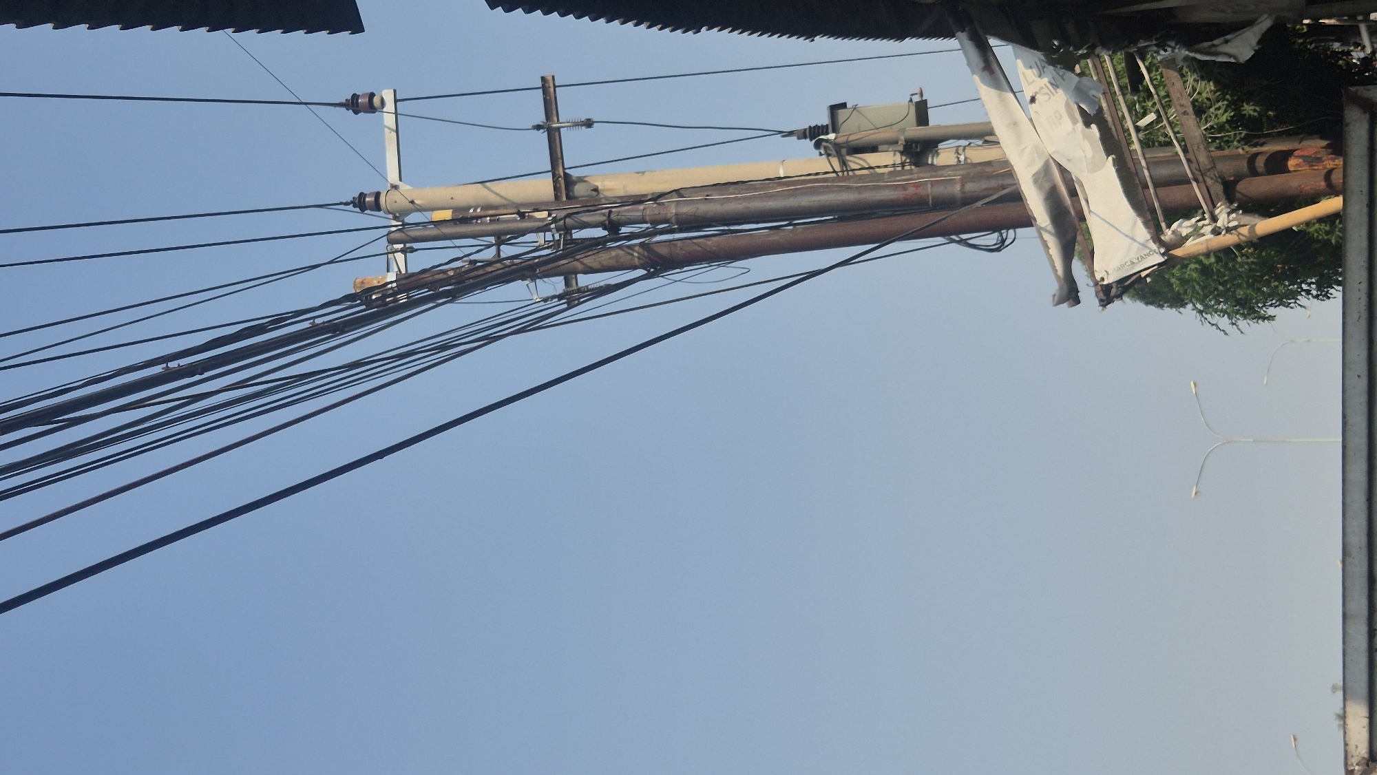 An outdoor, eye-level, vertical medium shot captures a cluster of utility poles against a clear, light blue sky. The poles, varying in color from off-white to rusty brown, are heavily laden with a dense network of black wires. One of the poles has a grey transformer box attached to it, and a small, light brown object, possibly a bird, is perched on one of the wires. To the right, the corrugated metal roof of a building is visible, as are the branches of a green tree. In the lower portion of the image, two streetlights with white, curved arms stand out against the sky, suggesting an urban setting. The overall impression is one of a densely wired urban environment in Karawang, Indonesia, under a bright, clear sky.