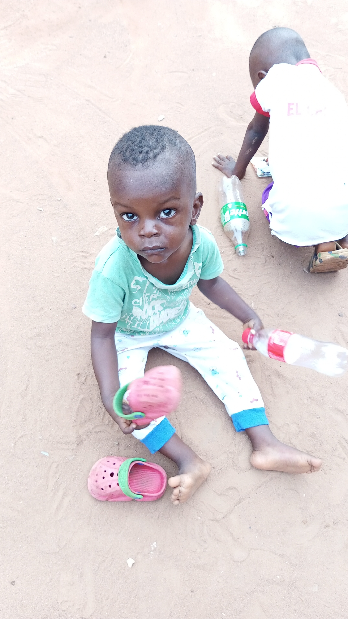 Two young children are in an outdoor environment in Onitsha, Nigeria. The ground is covered in light brown soil or sand. One child, sitting and facing the camera, has short black hair, is wearing a light green t-shirt, and patterned white pants with blue trim. They hold a pink plastic shoe with a green handle. Their left foot is bare, and another pink plastic shoe rests nearby. They hold a plastic water bottle in the other hand. Another child, visible from the shoulders down, stands in the background, wearing a white t-shirt with red trim and sandals. There is a plastic bottle lying on the ground near them.