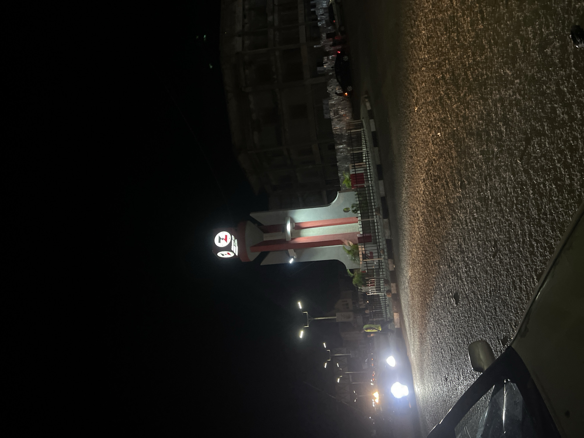 This image captures a nighttime urban scene at the Heritage Bank Roundabout (also known as Terminus Roundabout or Ahmadu Bello Way Roundabout) in Jos, Plateau State, Nigeria.
In the center of the frame stands a modern clock tower, illuminated with bright white light and accented with prominent red vertical stripes on a lighter grey base. The four clock faces on the upper section of the tower clearly display the stylized "HZ" logo of Heritage Bank. The tower is surrounded by a low black metal fence enclosing a small landscaped area with visible greenery in red planters.
The foreground features a dark, wet asphalt road, reflecting the bright white headlights of an approaching vehicle on the left side. The road surface exhibits a speckled texture, possibly from recent rainfall or poor surface quality, with scattered lighter reflections. A white and black striped curb marks the edge of the central island.
In the mid-ground, multiple streetlights with bright, rectangular fixtures illuminate the road. To the left of the clock tower, a person is visible wearing a light-colored (possibly yellow or lime green) reflective vest, standing near the roadside. Various other vehicles, identified by their illuminated headlights and taillights, are present in the background traffic.
To the right, behind the clock tower, a multi-story building with multiple window openings is discernible in the dim ambient light, appearing partially constructed or in a state of disrepair. Below this building, along the roadside, a large quantity of stacked clear plastic items, possibly crates or bottled goods, is visible. The sky above is completely dark, indicative of nighttime.