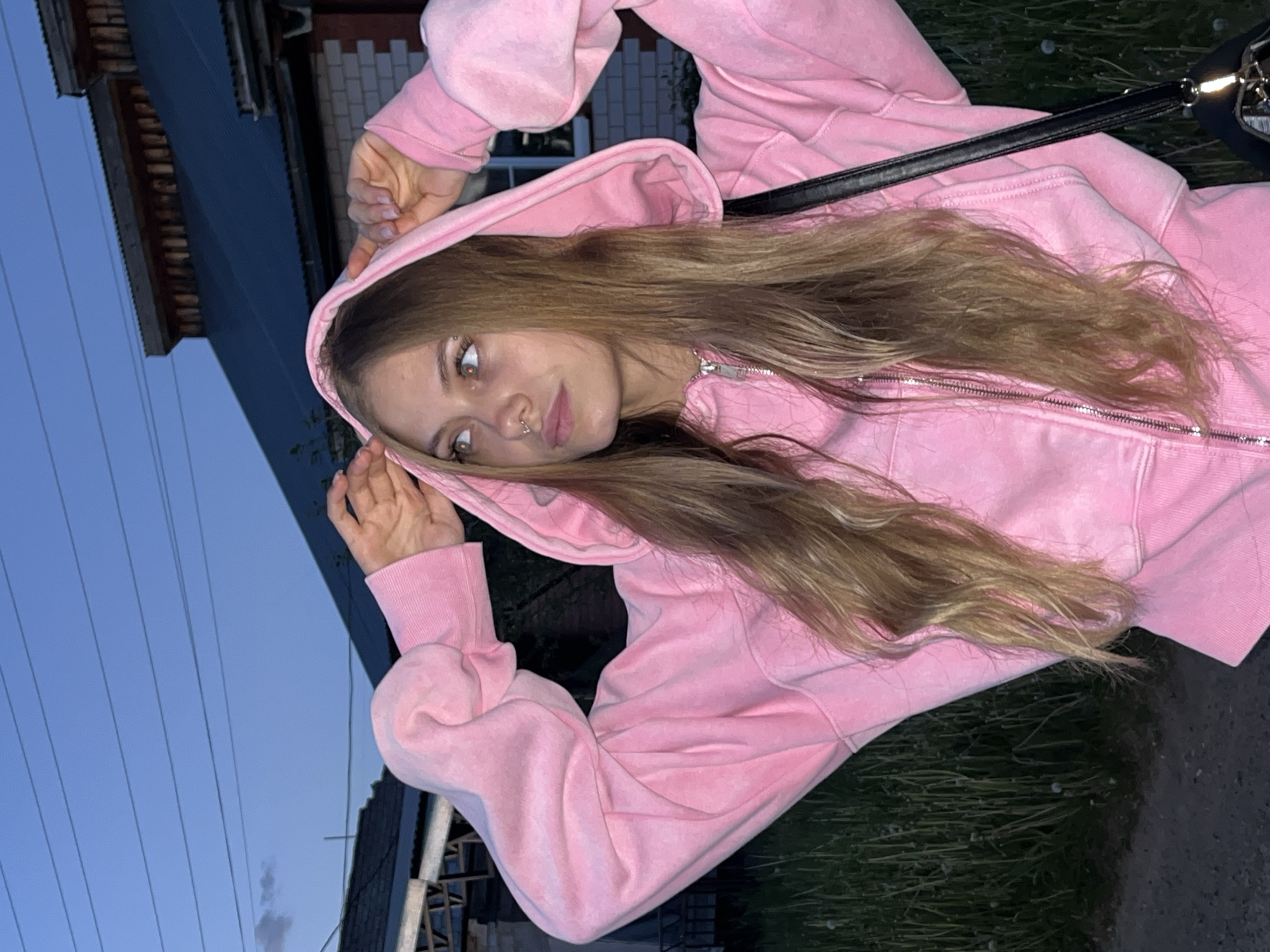 A young woman with long, blonde hair is wearing a pink hooded sweatshirt and standing outdoors. Her hands are near her face, possibly adjusting the hood. She has a nose piercing and appears to be looking at the camera. A black shoulder bag strap is visible across her shoulder. The background shows a building with brick accents, utility poles, and some greenery, suggesting an urban or suburban setting at dusk or twilight. Without geotagging information, it is not possible to confirm the precise location is Фоки, Russia, but the image provides no information to dispute it.