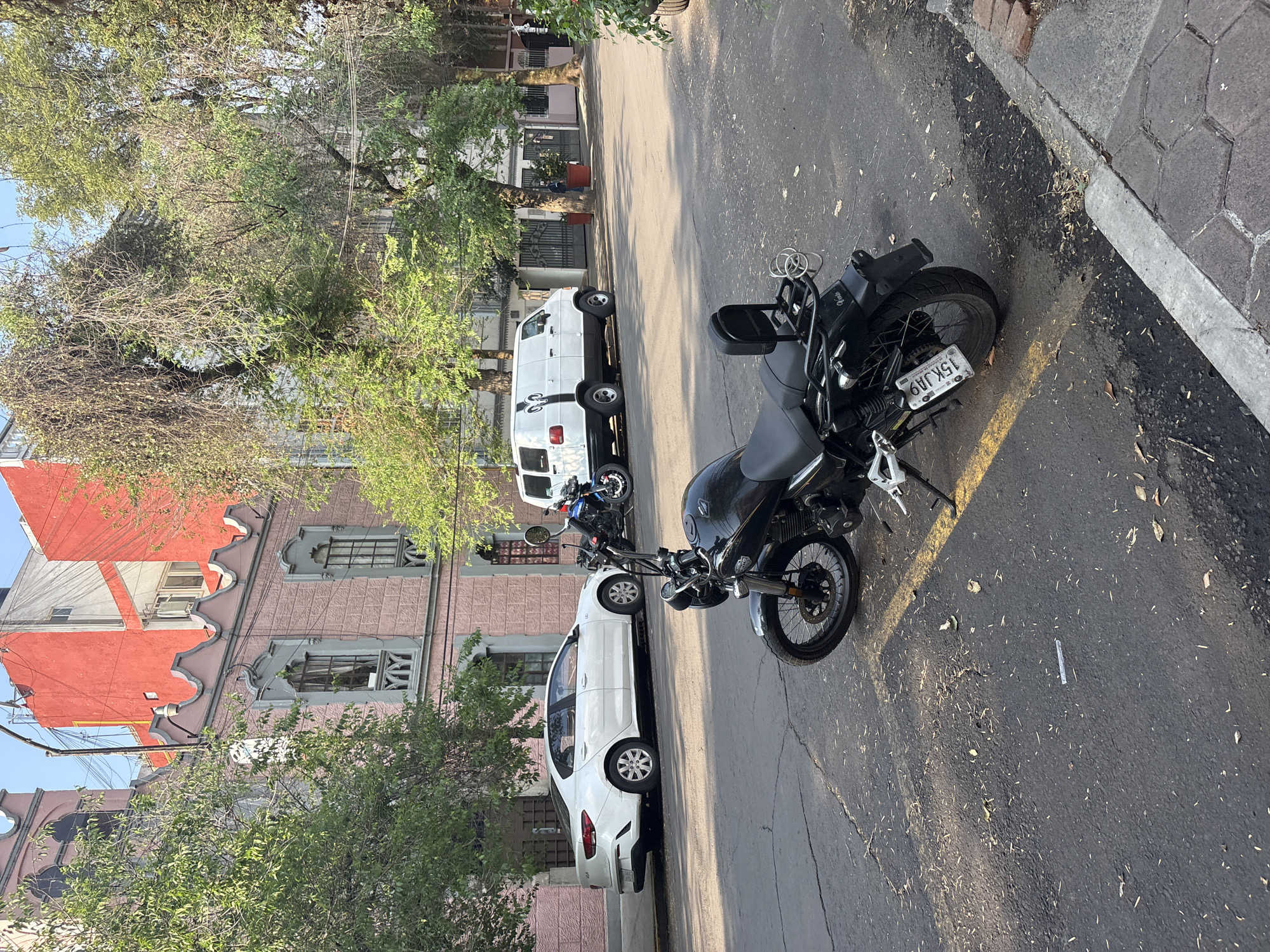 The image captures a street scene in Mexico City, Mexico. A black motorcycle is parked on the right side of the frame, partially obstructing the view of a white sedan parked alongside the curb on the left. Behind the sedan, a white van is also parked. A second motorcycle is visible behind the white car. The street is paved and marked with a yellow line. Buildings with pink and red facades, adorned with ornate architectural details, line the left side of the street. Trees with green foliage are interspersed along the sidewalk. The lighting suggests it is daytime.
