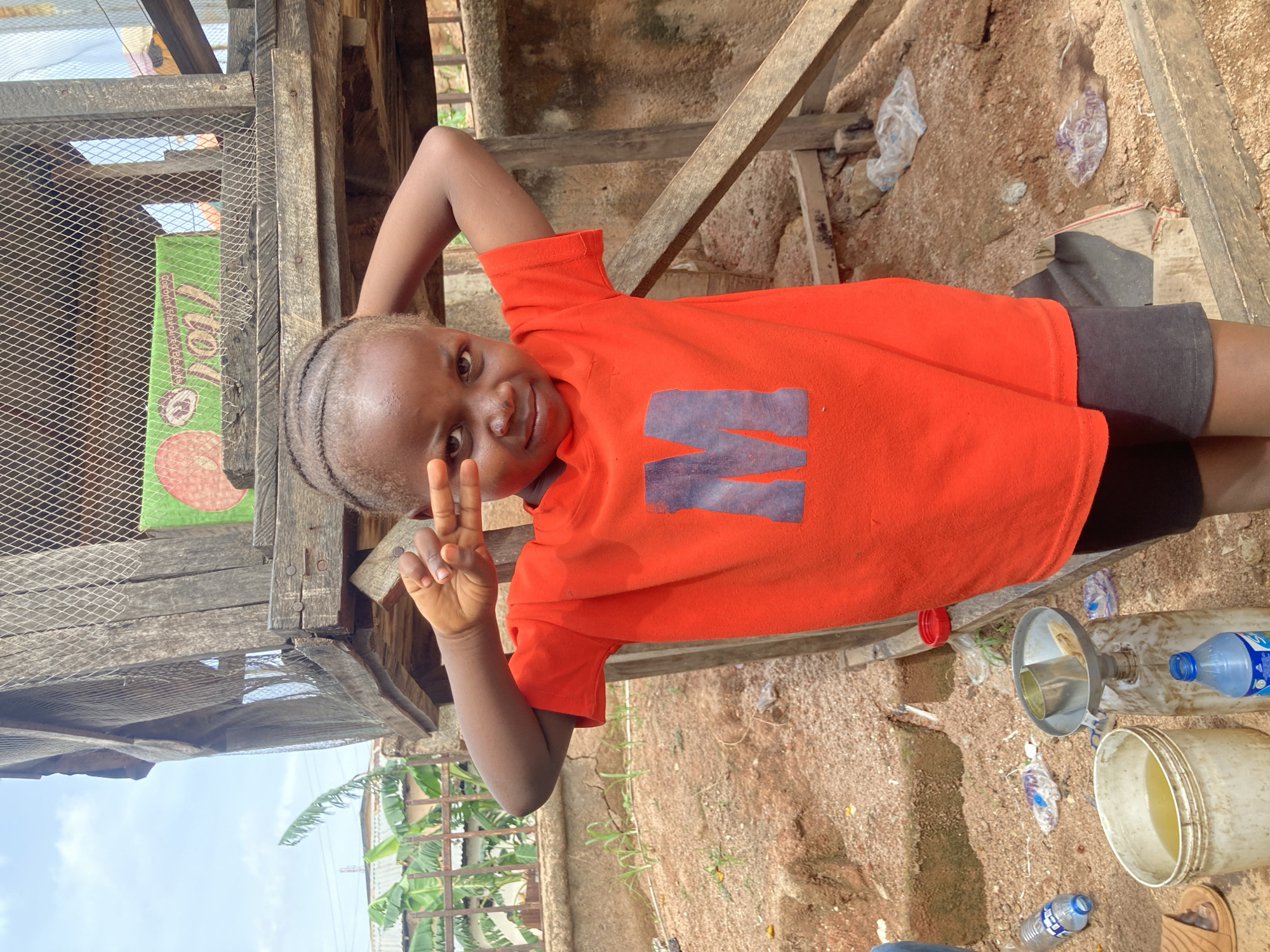 In Gwong, Nigeria, a young child stands in an outdoor area with a rough, sandy ground and a rustic wooden structure behind them. The child, wearing an orange t-shirt with a large blue letter "M" on it and dark shorts, poses with their hands near their face, forming a peace sign with one hand. The background includes elements of a rural or semi-urban setting: banana plants and what appears to be a corrugated metal roof of a dwelling are visible to the left. In front of the child, on the ground, are various items including plastic bottles, a white bucket with yellow liquid, and a funnel attached to a plastic container. The overall scene suggests a simple, everyday environment.