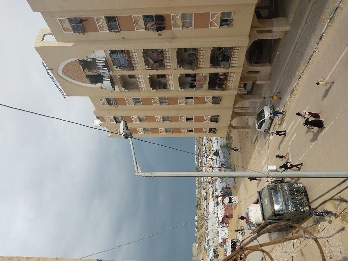 A multi-story building stands adjacent to a street with parked cars and pedestrians. Beyond the building, a dense settlement of smaller structures is visible. A utility pole with numerous wires extends into the sky, which is partly cloudy. The image is rotated, giving a skewed perspective of the scene.