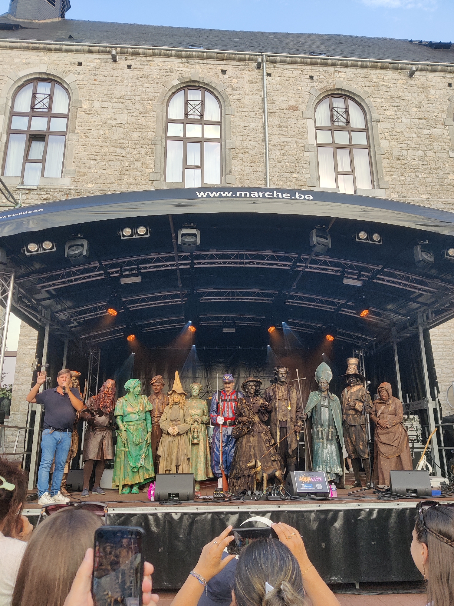 The image shows an outdoor stage in front of a stone building with arched windows. A man in a dark blue polo shirt and jeans is speaking into a microphone on the left side of the stage, his right hand raised. Behind him, a group of performers are standing on the stage. Most of them are dressed in elaborate costumes and appear to be painted or costumed to look like statues or figures from historical periods. The performer closest to the man speaking is dressed as a bearded man in dark armor. To his right, a performer is dressed in green, appearing as a female figure. Further to the right, several other performers are dressed in various historical costumes, including one in a tall hat and another in a bishop's attire. The stage is lit with orange spotlights. The building behind the stage has the text "www.marche.be" visible across the awning. In the foreground, several people are holding up their phones to take pictures of the performers. The sky is a clear blue, suggesting it is daytime. The scene appears to be a public event or performance taking place in the city of Marche-en-Famenne, Belgium, as indicated by the website address on the awning.