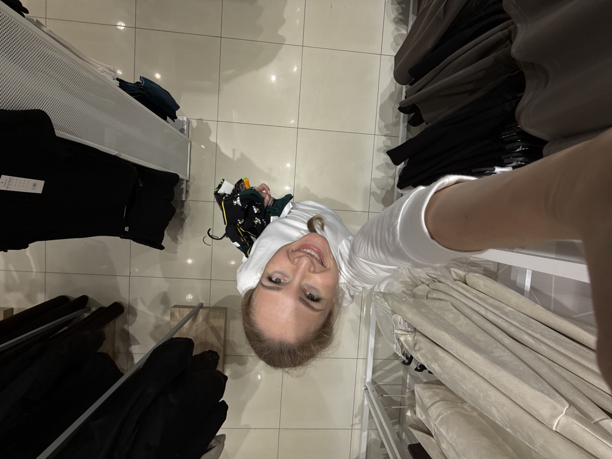 A woman is taking a selfie in what appears to be a clothing store. She is standing between two racks of clothing, one with dark-colored garments and the other with light-colored garments. There is a bag on the floor near the woman, and the floor is tiled. There is no visible information to establish the location in Chaikovsky, Russia.