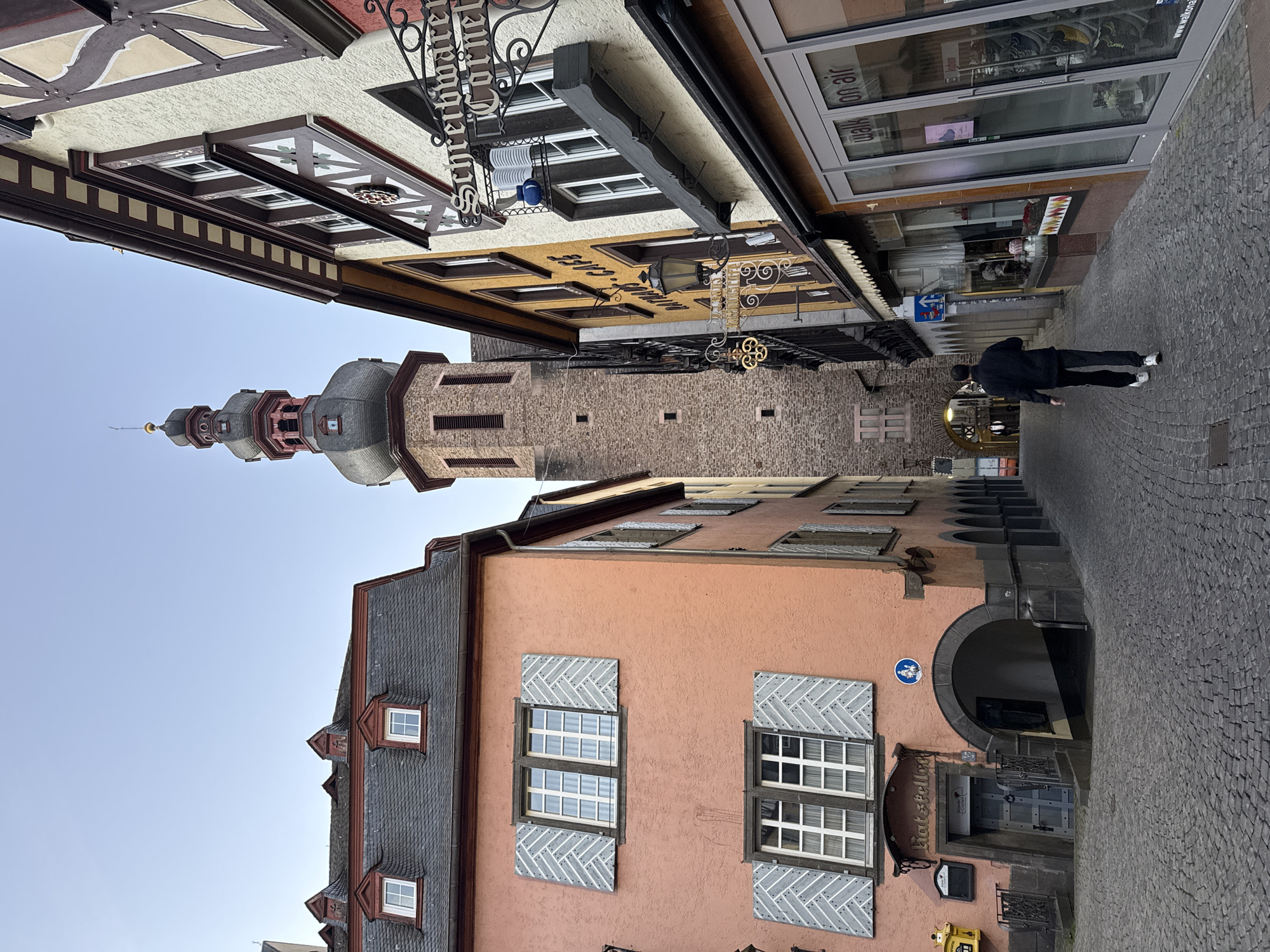 A street view in Cochem, Germany, depicts a person walking along a cobblestone street with tall buildings lining both sides. The buildings feature varied architectural styles, some with shuttered windows and others with decorative signage. A prominent medieval tower rises in the background, indicating the historic nature of the town. The sky is clear, suggesting daylight. The scene is primarily urban with a blend of residential and commercial structures.