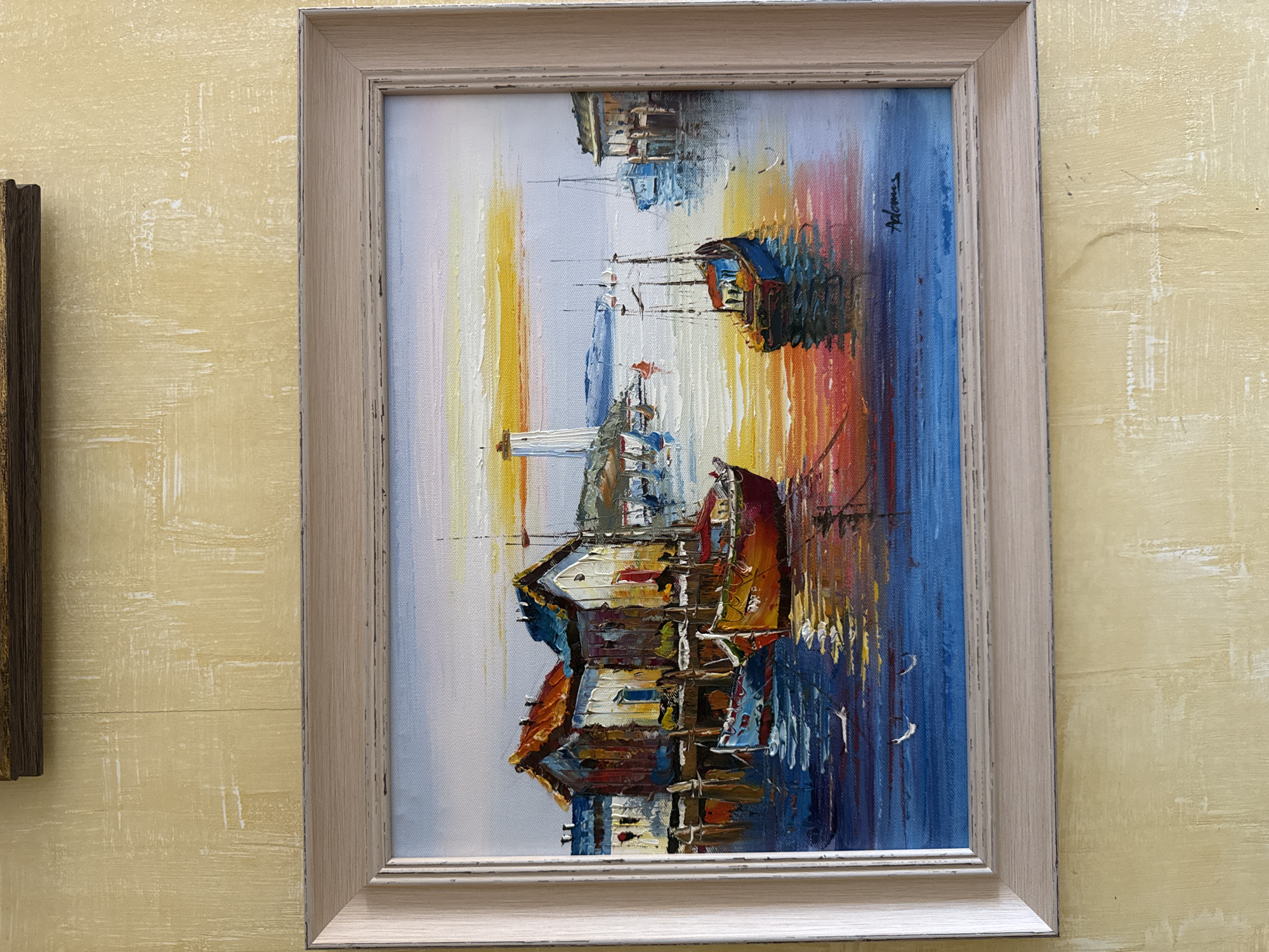 This is an oil painting of a harbor scene at sunset, rendered in a textured impasto style. The foreground features several boats moored in the water, with their reflections shimmering on the surface. Behind the boats, a collection of weathered buildings with colorful roofs line a dock. In the distance, a lighthouse stands tall against a vibrant sky painted with hues of orange, yellow, and pink, characteristic of a sunset. The overall impression is a lively and colorful depiction of a coastal town. The provided image does not contain any information or visual cues that would allow for the identification of the location as Yeysk, Russia.