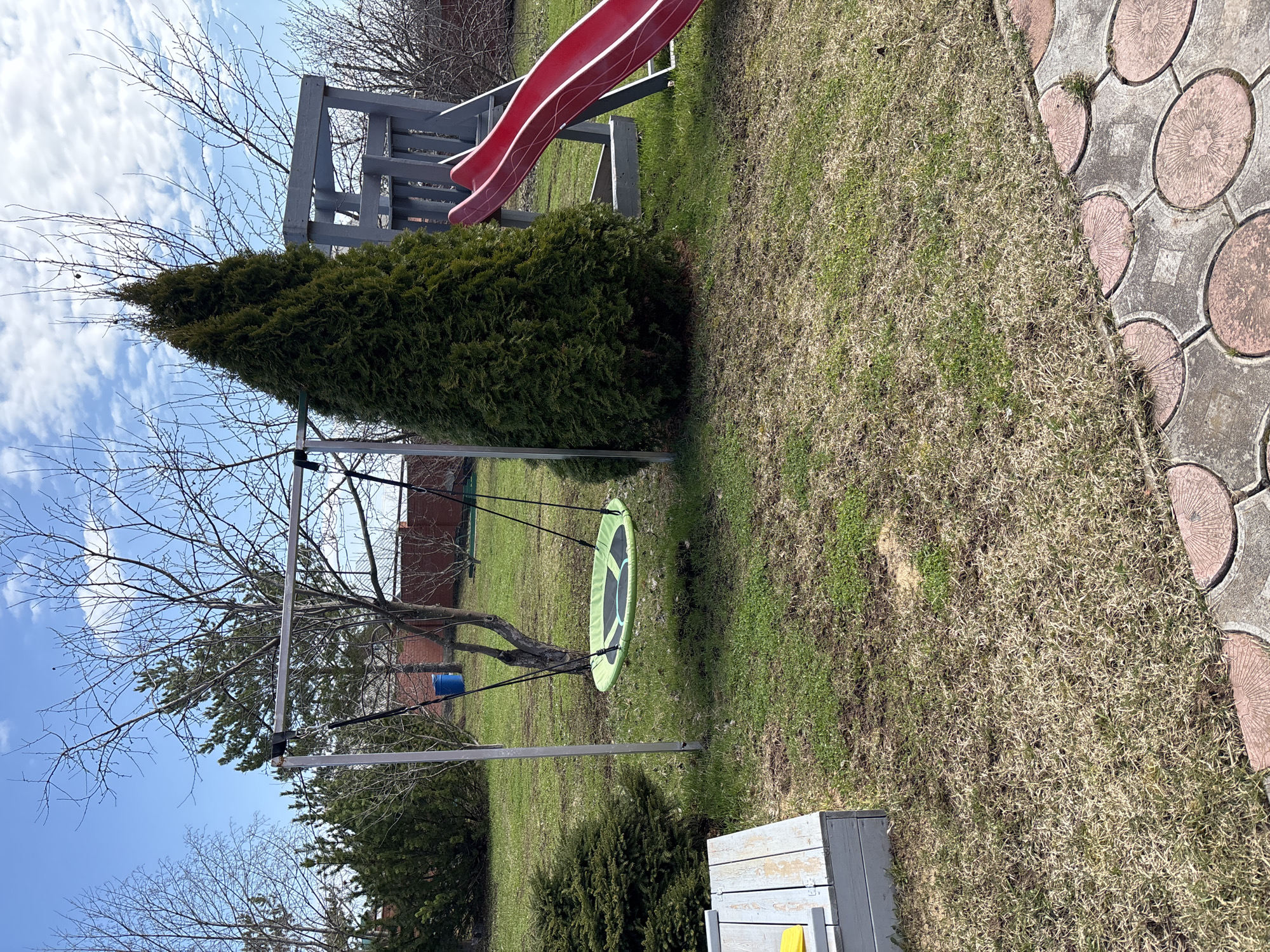The image depicts a backyard scene in Ферма, Russia, featuring a swing set, a slide, and various landscaping elements. The swing set is silver and supports a green circular swing. A leafless tree grows through the swing set structure. A red slide stands in the background near a conical evergreen bush. The yard has grass that is a mix of green and brown, suggesting early spring or late fall. A paved walkway is visible on the right side of the image. A white wooden box is in the lower right corner. The sky is blue with scattered clouds.