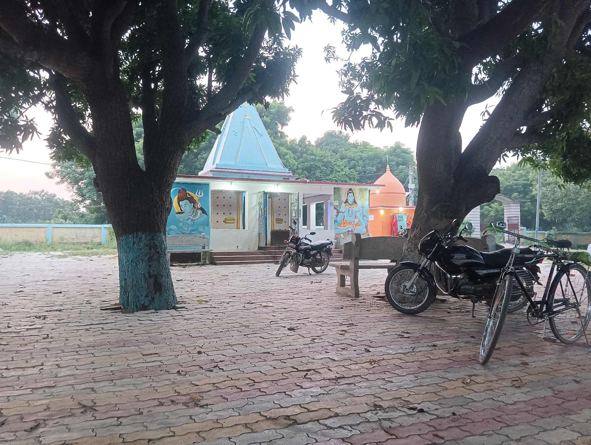 In the city of Tilkar, India, a temple complex is depicted with a paved courtyard. The temple features a prominent light blue pyramid-shaped spire and an orange dome. Two large trees with dense foliage frame the scene, one on the left with its trunk painted blue at the base, and another on the right. Murals of a deity, possibly Lord Shiva, are painted on the temple walls. Parked in the courtyard are two motorcycles and a bicycle. A concrete bench is visible near the tree on the right. The ground is paved with interlocking, multi-colored bricks. The environment appears to be outdoors, possibly in the early morning or late evening, given the soft light.