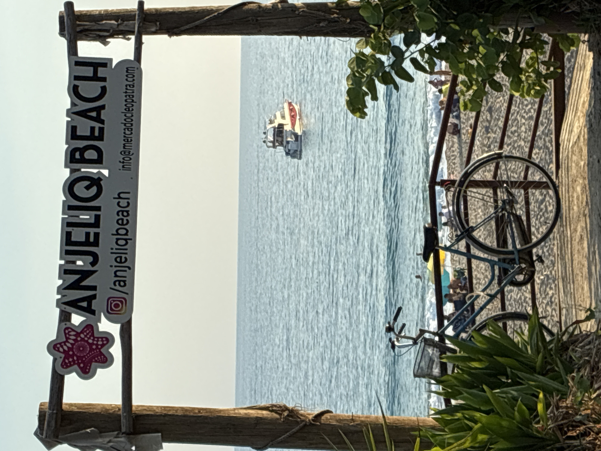 The image captures a view of Anjeliq Beach in Alanya, Turkey, from a wooden archway. A sign above the arch reads "ANJELIQ BEACH" with an Instagram logo and "@anjeliqbeach" on the left, and "info@mercadocleopatra.com" on the right. A bicycle with a basket is parked in the foreground, leaning against a wooden railing that separates the pathway from the sandy beach. Beyond the railing, people are seen relaxing on the beach under umbrellas, and the blue sea stretches to the horizon. A white and red boat is visible on the water. Lush green plants frame the right side of the scene.