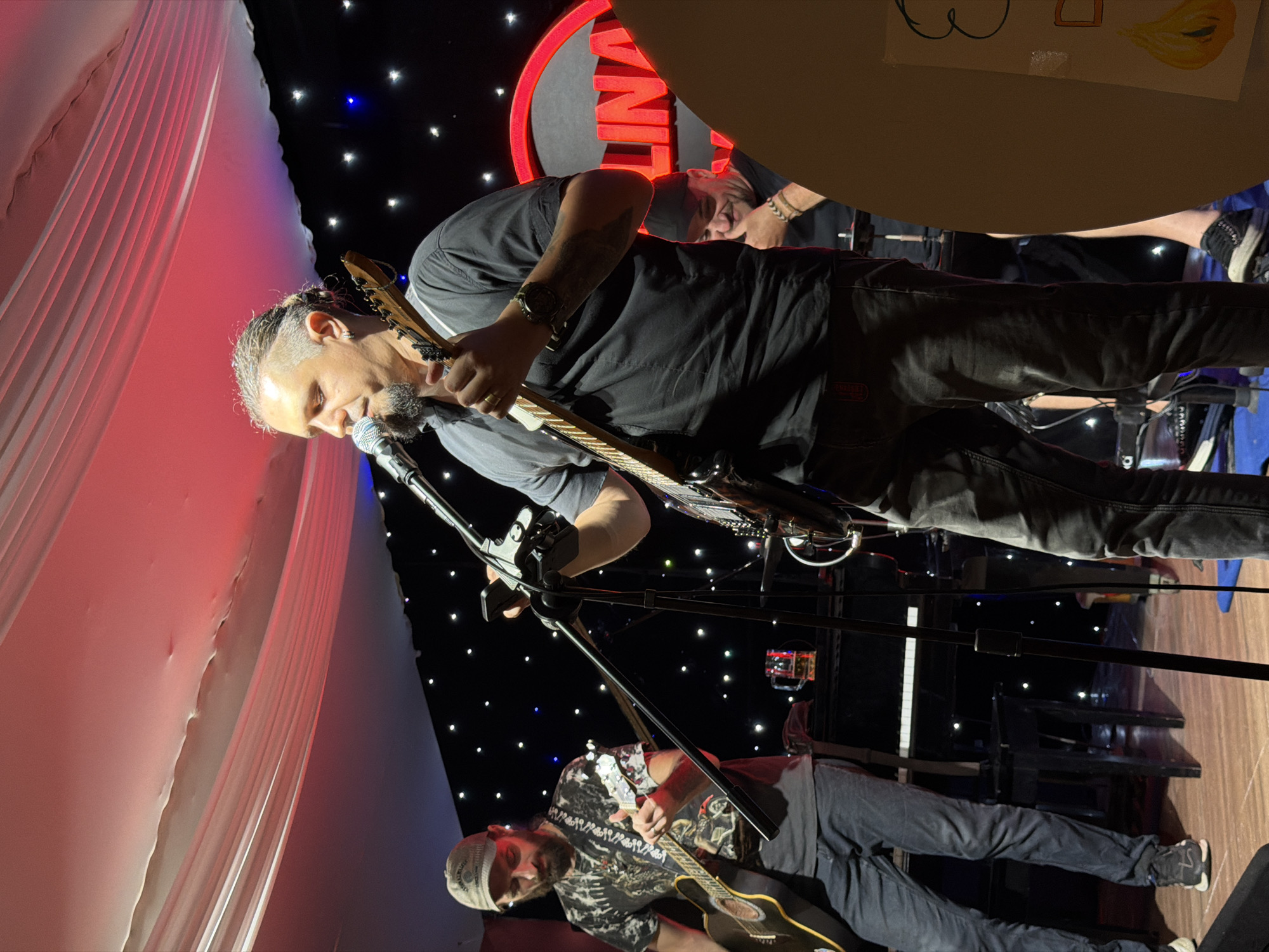 In the city of Nha Trang Ward, Vietnam, a live music performance is underway on a stage. Three men are visible, all engaged in playing instruments or singing. The central figure, a man with his hair braided and pulled back, sings into a microphone while holding an electric guitar. To his left, another man, wearing a baseball cap and a patterned t-shirt, strums an acoustic guitar. Behind the central performer, a third man in a black cap and t-shirt smiles and rests his chin on his hand, appearing to be part of the band or audience.
The stage is adorned with draped fabric creating a canopy overhead, illuminated by red and blue lights that cast a vibrant glow. A backdrop of black fabric is punctuated by numerous small, twinkling lights, resembling stars. A piano is visible behind the acoustic guitarist, and a neon sign with the word "LINA" in large red letters is prominently displayed in the background. The overall ambiance suggests a lively and intimate concert setting.