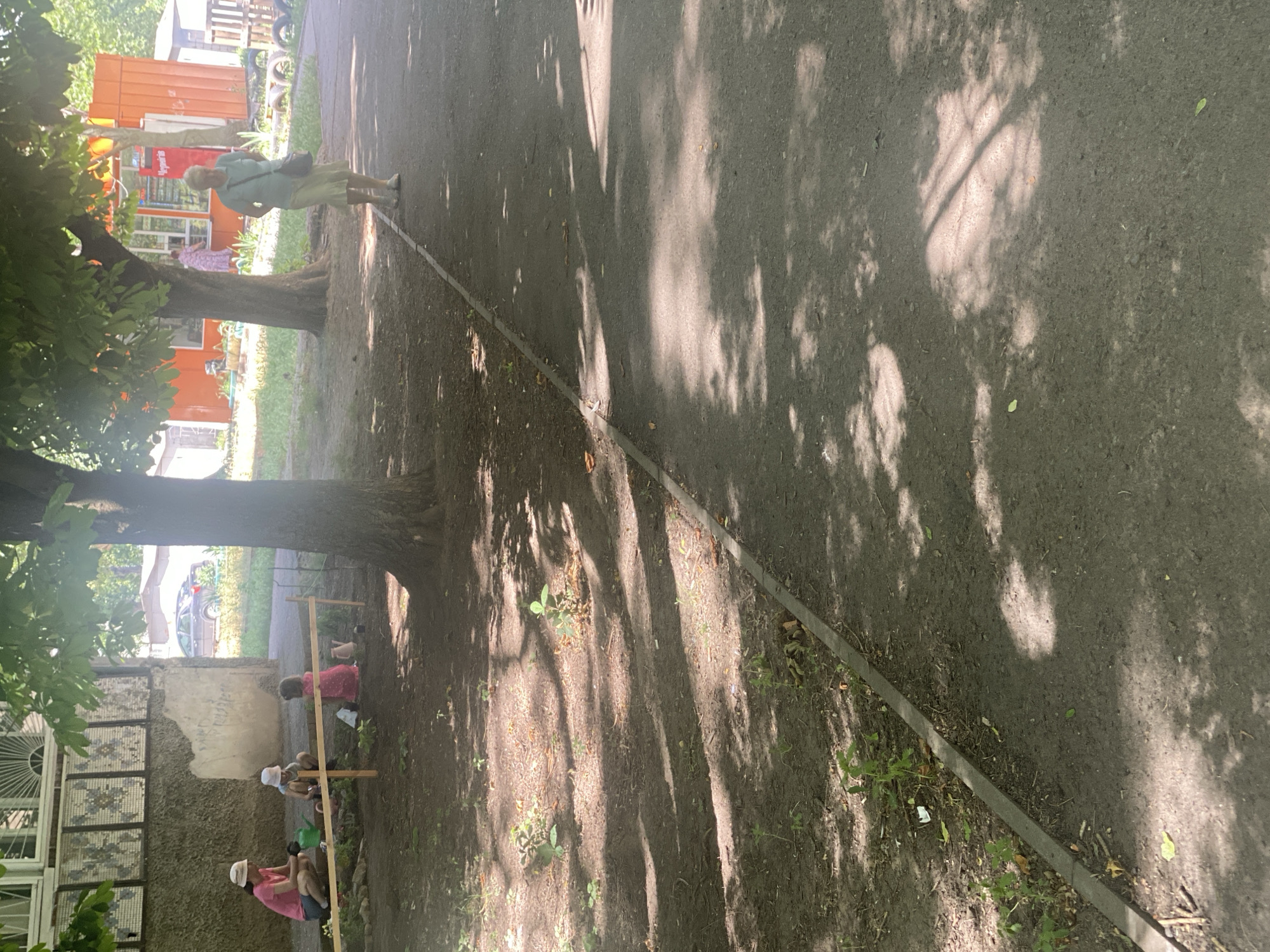 In Odesa, Ukraine, an outdoor scene unfolds under dappled sunlight, likely a public space adjacent to a building. In the foreground, a section of paved pathway runs diagonally, bordered by a dirt area. This dirt area is sparsely populated with small plants and scattered leaves. The pathway is partially covered in shadows cast by overhead trees.
To the left, a group of three individuals are present in the dirt area. A woman wearing a pink t-shirt and a light-colored hat crouches near a shallow wooden enclosure, holding a green watering can. Two children are within or near this enclosure. One child, also wearing a light-colored hat, sits down. Another child, dressed in a pink dress, is seated. A small pink watering can is visible nearby.
Further in the background, an orange kiosk-like structure stands, suggesting a small shop or vendor. A woman with light-colored hair, wearing a light blue top and a light skirt, walks on the paved area towards the kiosk. The building behind the initial scene features a textured concrete wall with decorative mosaic panels and a window with a metal grille. A car is parked in the distance. The overall environment appears to be a casual, outdoor gathering space, possibly a park or a courtyard.