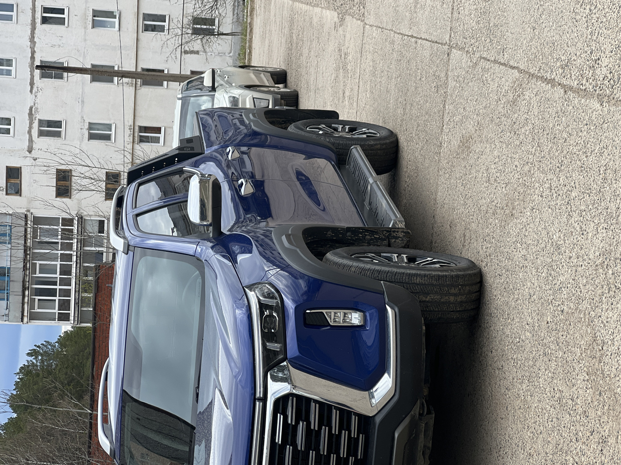 A dark blue pickup truck is prominently featured, parked on what appears to be a paved street or parking area in Noviy, Russia. The truck, facing right from the viewer's perspective, displays a modern design with a large chrome grille, distinct LED headlights, side steps, roof rails, and chrome side mirrors. Its tires show good tread. Behind it, a lighter-colored, possibly silver or grey, pickup truck is partially visible.
In the background, a multi-story building with numerous rectangular windows occupies a significant portion of the frame. The building's facade is light-colored, possibly concrete or stucco, and shows signs of wear and texture. Bare tree branches are visible between the trucks and the building, suggesting late winter or early spring. Further in the distance, hints of green trees and another building can be seen. The ground is a light-colored, textured concrete or asphalt surface.
The scene is illuminated by bright, diffused daylight, indicating either an overcast sky or an indirect light source, as no harsh shadows are cast. There are no people visible, and the trucks appear to be stationary. No legible text or brand names are clearly visible on the vehicles or the building.