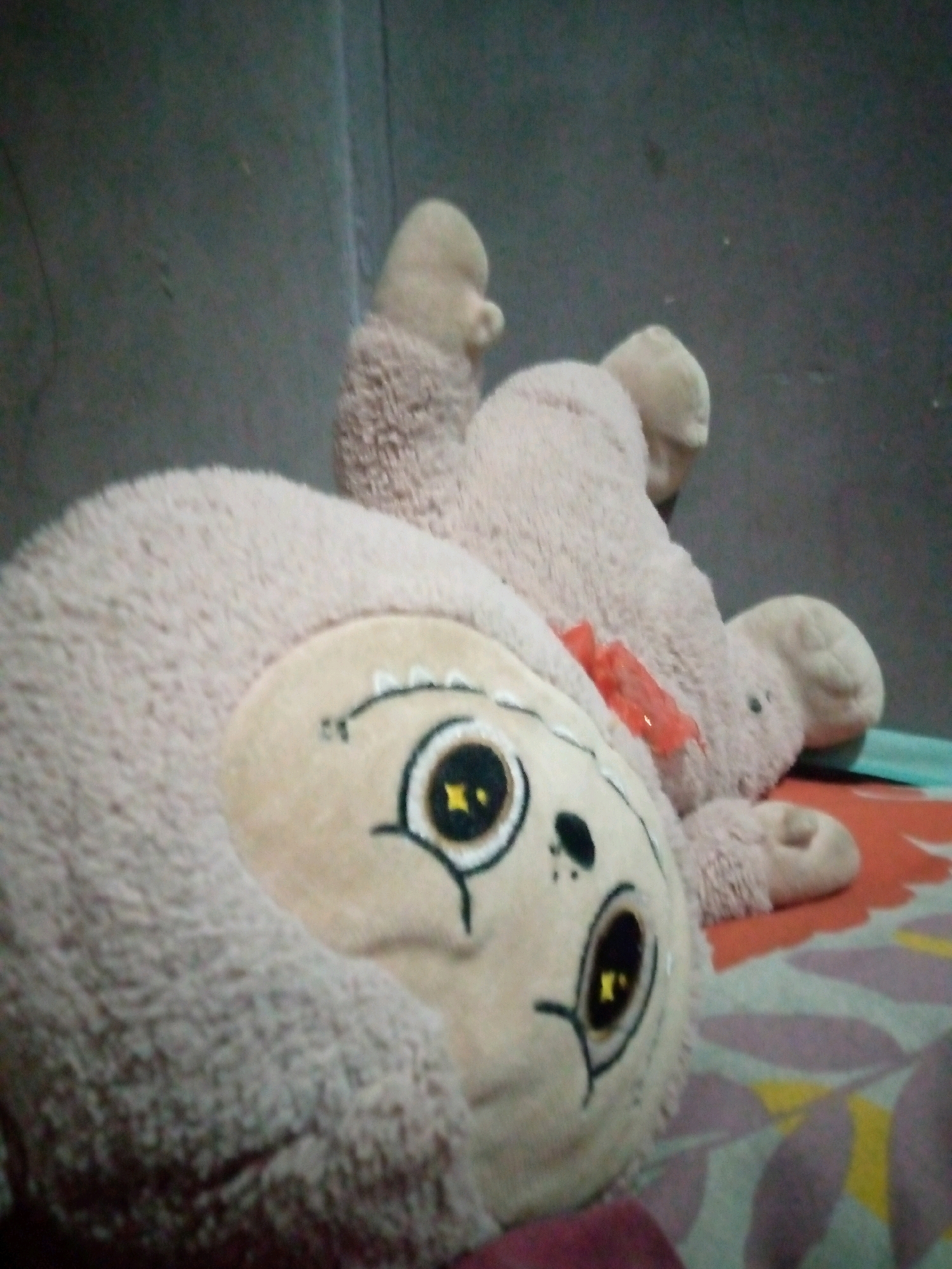 A light brown, fuzzy stuffed sloth toy lies on its back on a bed with a colorful patterned blanket. The sloth's face is visible, featuring large, round eyes with yellow irises, black pupils, and outlined in black. Above the eyes are short black lines representing eyebrows, and a few black dots suggest freckles or a nose. A small red ribbon is tied around the sloth's neck. The background appears to be a textured, gray wall. The scene is indoors.
