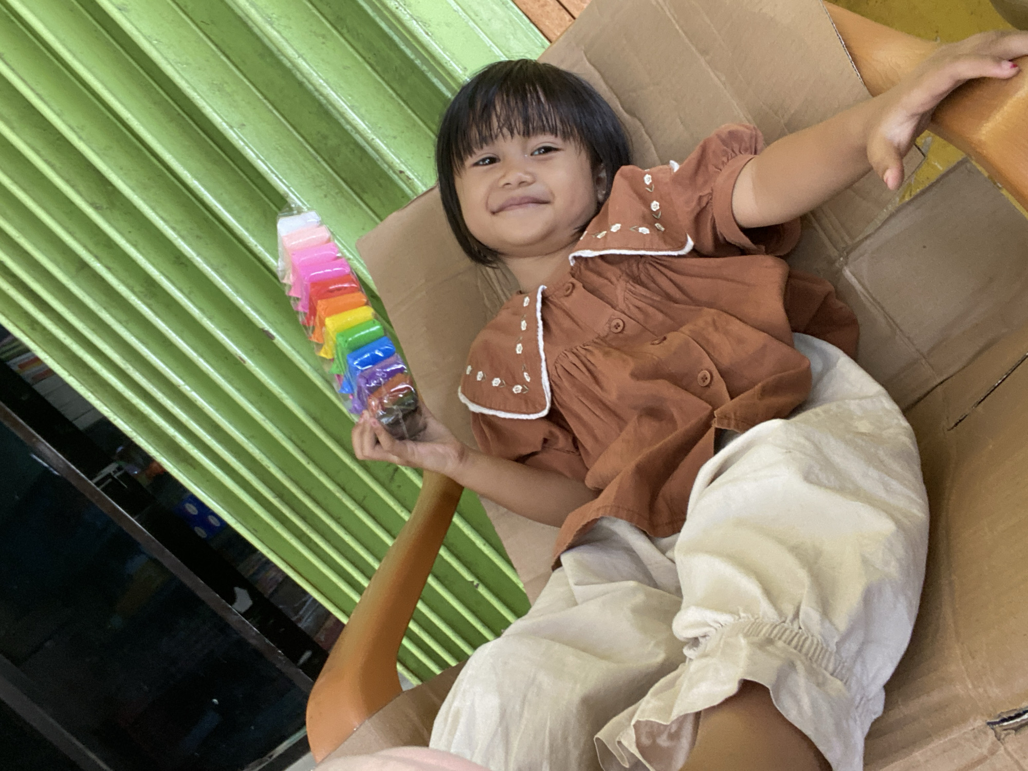 A young girl lies on a cardboard box, holding a pack of rainbow-colored modeling clay. She is wearing a brown shirt with a white collar adorned with floral embroidery and light-colored pants. The background features a green corrugated metal wall. The scene appears to be an indoor or semi-outdoor space, potentially a workshop or a storage area, given the presence of cardboard and industrial-looking blinds.