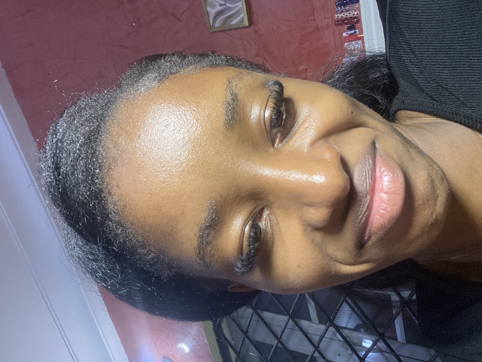 A close-up, low-angle selfie shows a young Black woman with dark hair styled upwards, revealing wisps of grey at her hairline, possibly indicating premature greying. She has noticeable eyelashes, smooth, shiny skin with a slight sheen, and her lips are slightly parted in a subtle smile. The background is a textured reddish-pink wall, with a barred window on the left and a framed picture on the right. The overall impression is of a casual, indoor setting. No specific location in Jos, Nigeria is identifiable.