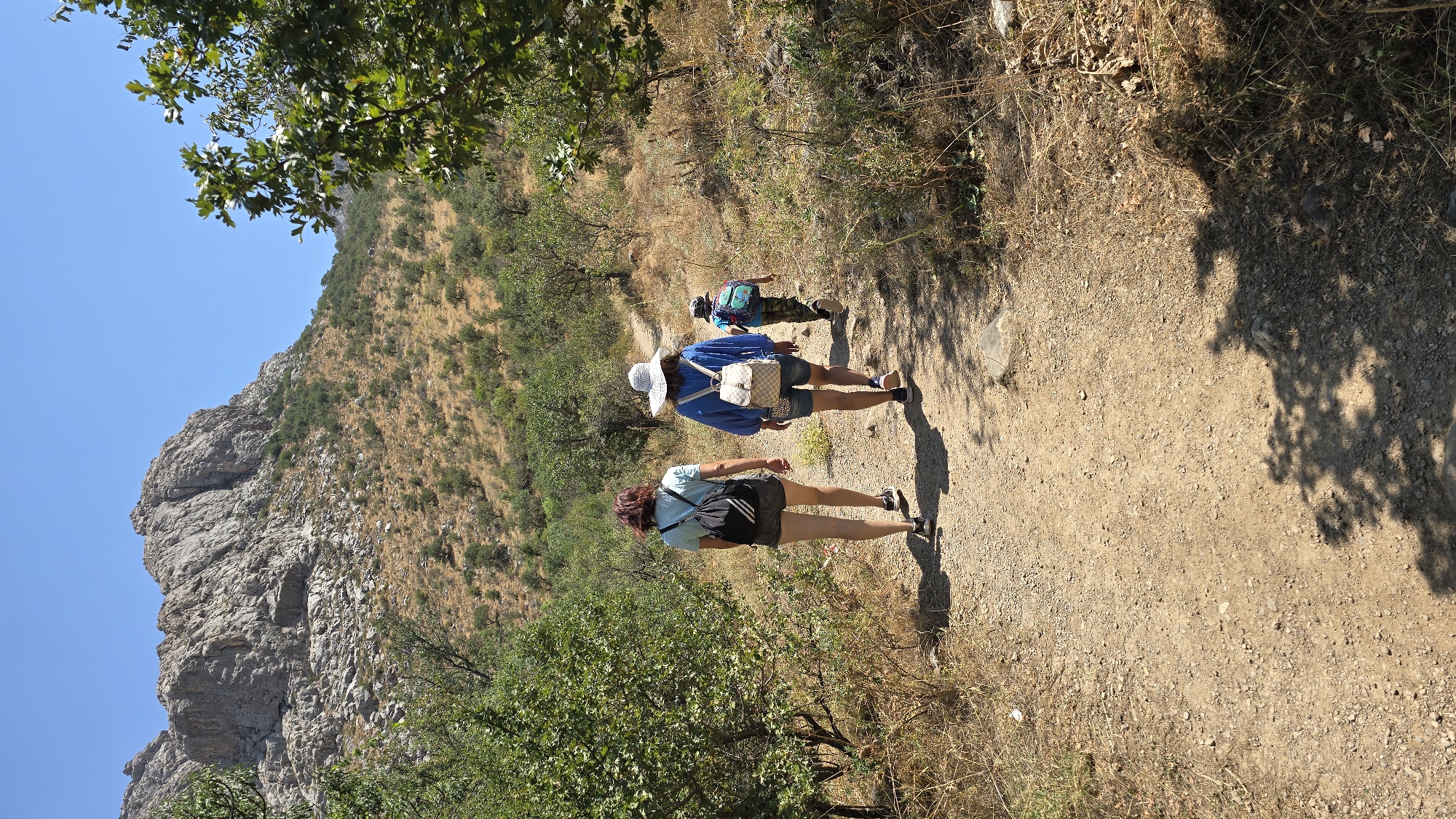 In the city of Terekty, Kazakhstan, three individuals are captured walking along a dirt path that winds through a dry, hilly landscape. The path is surrounded by sparse, dry vegetation and scattered trees, with a prominent rocky mountain dominating the background under a clear blue sky. The group consists of two adults and one child, all walking away from the camera. The adult in the lead wears a light blue t-shirt, dark shorts, and a black backpack. Behind her, another adult is dressed in a blue long-sleeved shirt, light-colored shorts, and a straw hat, carrying a light-colored backpack. The child, in the rear, wears a camouflage-patterned shirt and shorts and a colorful backpack. All three appear to be on a hike, with the environment suggesting a warm, sunny day.