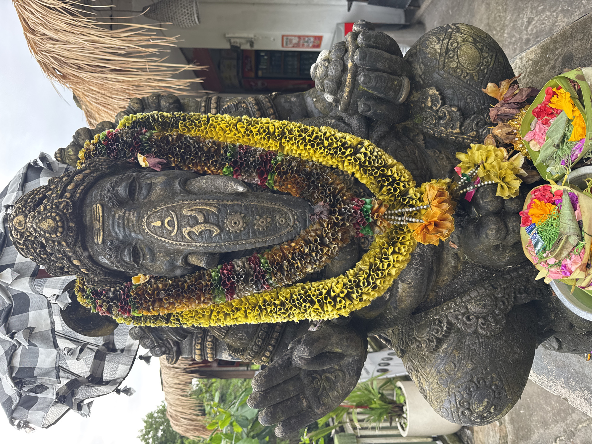 A large, weathered stone statue, likely depicting the Hindu deity Ganesha, is the central subject of the image, presented in a portrait orientation. The statue is made of dark, textured stone, possibly volcanic rock, with intricate carvings highlighted by gold-colored accents on its crown and body.
Around the statue's neck and chest are multiple garlands made of artificial or dried flowers, predominantly bright yellow and darker hues of red and green, creating a vibrant contrast against the dark stone. A prominent "Om" (ॐ) symbol is carved onto the statue's chest/belly. A traditional Balinese black and white checkered cloth, known as "poleng," is draped over the statue's left shoulder and arm.
To the right of the statue, several fresh offerings are placed. These include small, square woven palm-leaf baskets, known as "canang sari," filled with colorful fresh flowers in shades of red, yellow, and pink, interspersed with green foliage. Some loose dried flowers are also visible near these offerings.
The setting appears to be outdoors, with a traditional thatched roof, possibly made of alang-alang grass, visible in the upper left background. Green foliage from potted plants can be seen in the lower right. The overall atmosphere suggests a serene, devotional space, likely a temple or shrine in Bali, Indonesia, consistent with Kuta Selatan. The soft, diffused lighting indicates it is daytime, possibly overcast. No human activity is directly depicted, but the fresh offerings imply recent religious rituals or acts of devotion.