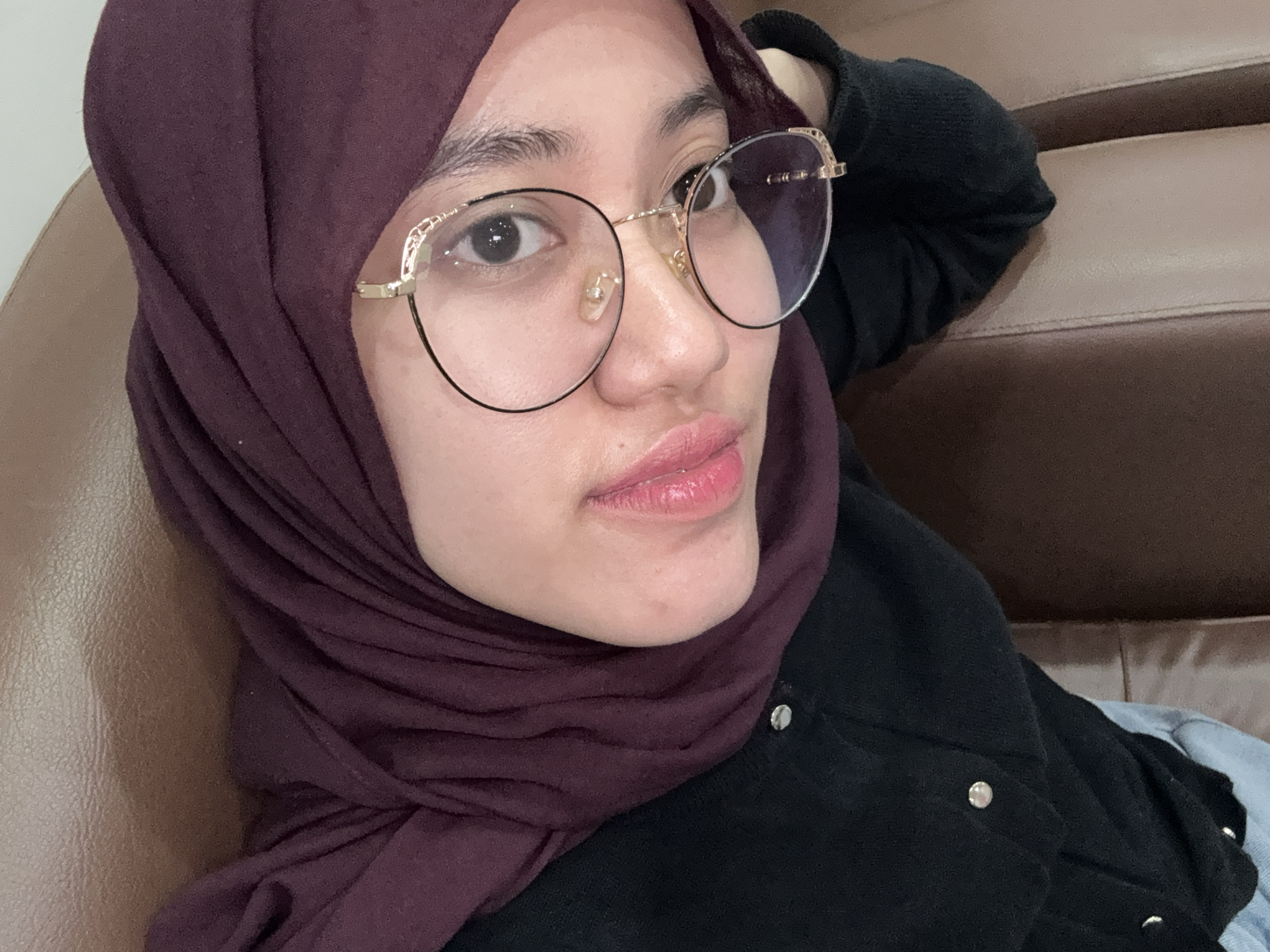 A close-up, chest-up portrait features a young woman of Asian descent, likely in Kaliwates, Indonesia, relaxing in an indoor setting. She is positioned on a brown leather or faux leather couch, which forms a significant part of the background.
The woman is wearing a dark maroon or deep purple hijab, gracefully draped around her head and neck. Her outfit includes a black top or jacket, detailed with prominent silver buttons visible on the front. A lighter, possibly blue, garment is partially seen on her right side. She sports large, round glasses with golden frames, notable for their decorative accents along the top outer edges.
Her gaze is directed slightly towards her right (the viewer's left), with her eyes open and clear. Her lips are subtly parted, displaying a natural, glossy appearance, suggesting the presence of lip gloss or natural hydration. Her expression appears relaxed and neutral, conveying a calm demeanor. The lighting is soft and even, consistent with an indoor environment, possibly illuminated by artificial light or indirect natural light. There is no visible text within the image. The scene suggests a casual and comfortable moment.