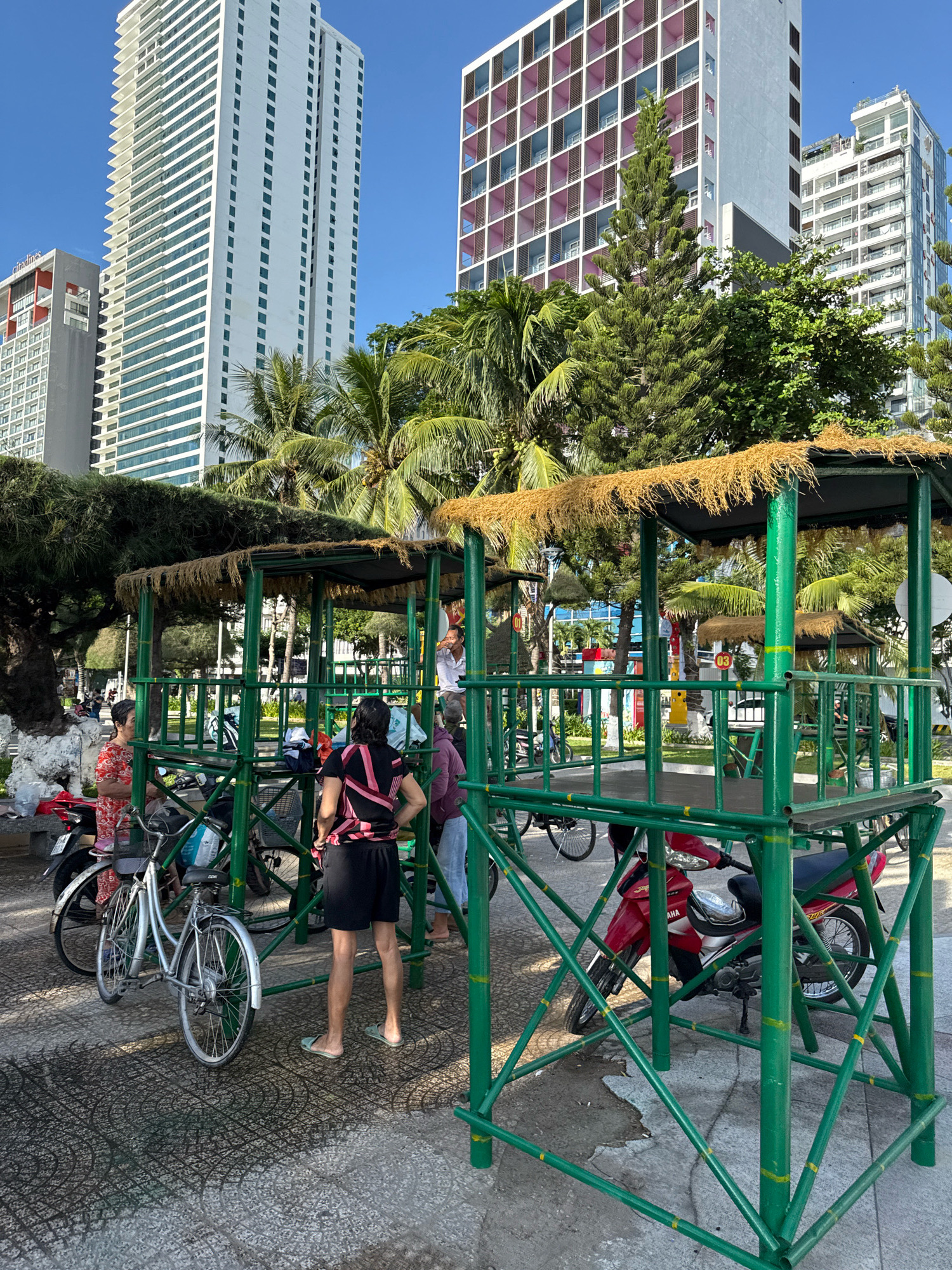 In Nha Trang, Vietnam, a street scene unfolds beneath a clear blue sky. Tall, modern buildings with patterned facades dominate the skyline, interspersed with lush palm trees and evergreens. In the foreground, a vibrant green metal structure, resembling a shaded pavilion or kiosk with a thatched roof, stands as a central focus. This structure appears to be a parking area or shelter, as several motorcycles and bicycles are parked within and around it. Two women, one in a red floral dress and the other in a black and pink striped top and black shorts, are standing near the structure. The woman in the floral dress is looking to her left, while the woman in the striped top faces away from the camera, her hands on her hips. Another person, partially visible, stands behind them. The ground is paved with patterned tiles, some appearing wet or discolored. The overall atmosphere suggests a warm, sunny day in a bustling urban coastal environment.