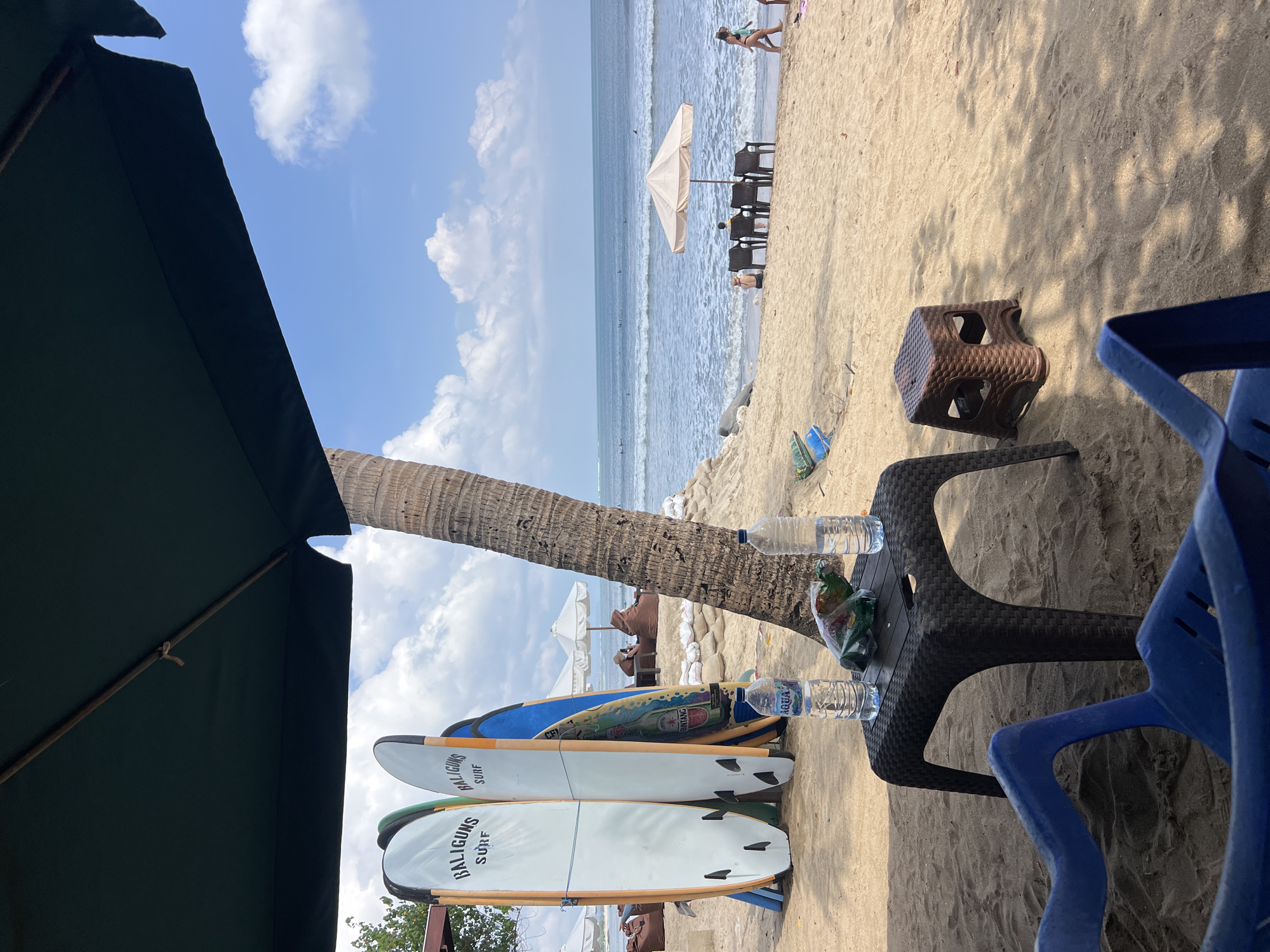 This image depicts a beach scene in Kuta, Indonesia, under a partly cloudy sky. In the foreground, two blue plastic chairs face away from the viewer, with a dark plastic table positioned between them. On the table, two bottles of Aqua water and a green plastic bag containing what appears to be snacks are placed. To the left of the table, three surfboards stand upright, two of which have "BALIGUNS SURF" written on them in black lettering. A palm tree trunk rises to the right of the surfboards. In the middle ground, several beach chairs with a tan umbrella are set up on the sand. Beyond the chairs, the ocean stretches to the horizon, with small waves breaking and a few surfers visible in the water. In the background, more beach umbrellas and sandbag structures are present. A person in a dark swimsuit is walking on the beach to the right of the umbrella. The overall environment is a tropical beach setting, likely a popular spot for surfing and relaxation.