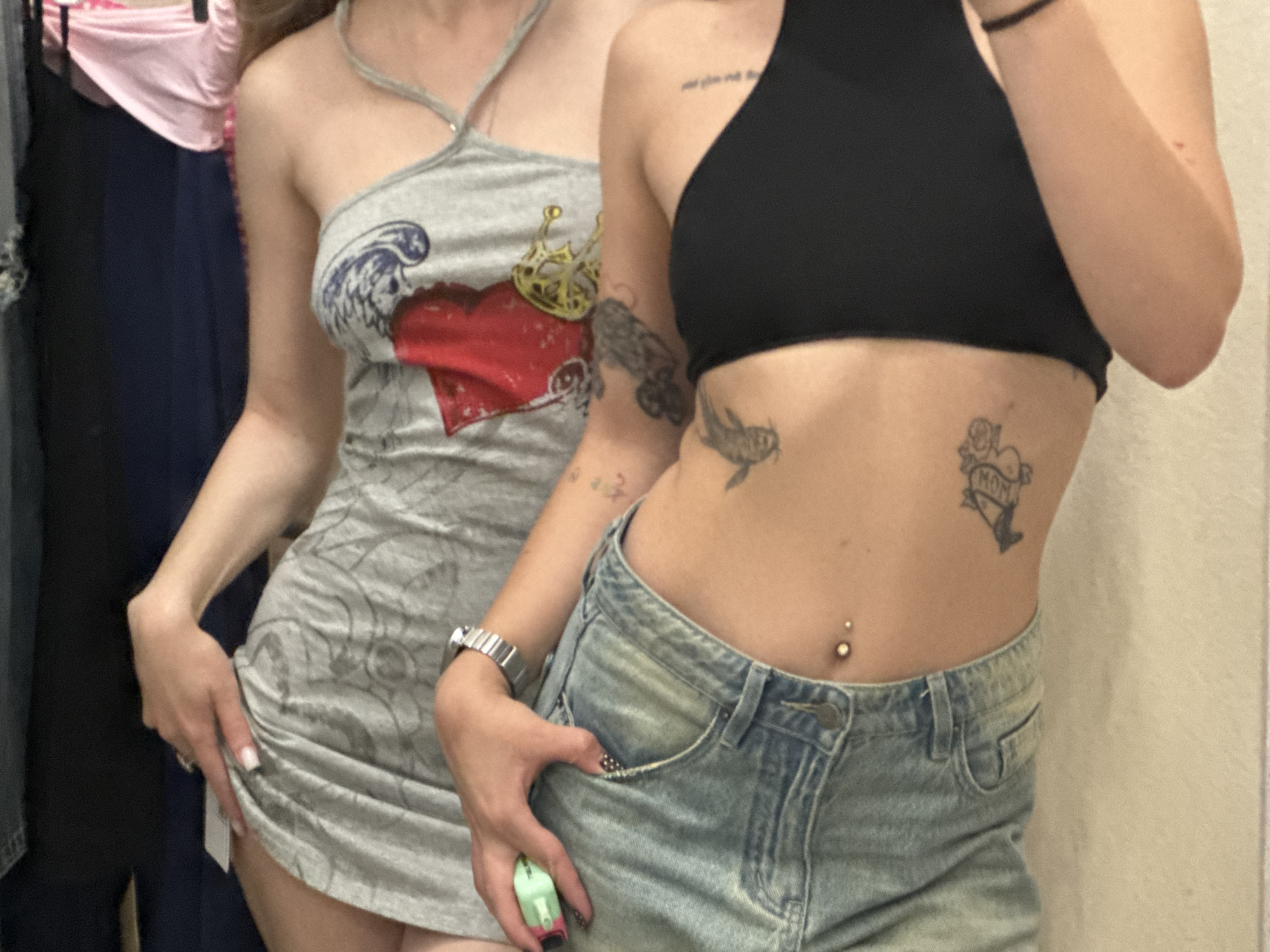 Two young women are depicted from the torso down, posing indoors, likely in a fitting room or retail environment in Moscow, Russia. The woman on the left wears a grey, ruched halter-neck mini-dress featuring a graphic design of a red heart topped with a golden crown and stylized blue waves. Her left hand rests on her hip, slightly pulling up the hem of her dress, revealing a small white tag. She has white nail polish and a silver ring.
The woman on the right wears a black one-shoulder crop top and light blue, distressed denim shorts. Her right arm is bent, with her hand resting casually in her front pocket. She wears a silver watch on her left wrist and has dark nail polish with white polka dots. A light green vaping device is held in her right hand.
Both women have visible tattoos. The woman on the right has several: partial text on her upper chest/shoulder, which appears to say "and give me peace," two distinct bird-like designs on her right ribcage, and another tattoo on her lower left ribcage that includes a rose and the word "MOM" within a ribbon design. She also has a belly button piercing with a gold barbell. The woman on the left has tattoos on her right arm, though their specific designs are unclear.
The background consists of blurred clothing hanging on racks, including pink and dark blue/black garments, and a plain, light-colored wall. The overall mood is casual and relaxed.