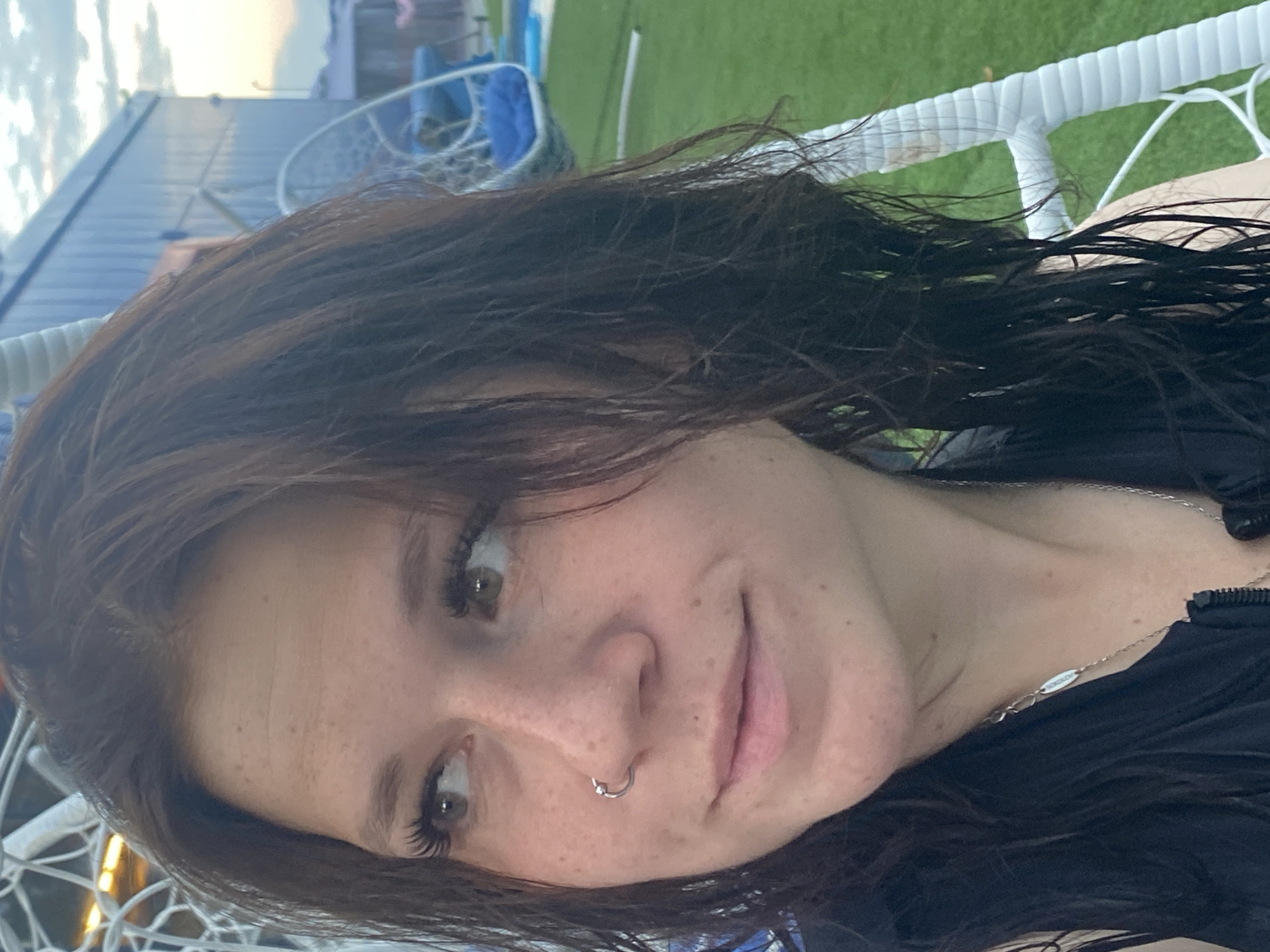 A close-up, head-and-shoulders shot captures a young woman with dark, wavy hair and a light complexion, adorned with freckles. Her gaze is directed to her left, and she has a silver hoop piercing in her nose. She is wearing a black top and a silver necklace. The background is blurred, but suggests an outdoor setting with a blue building and green artificial turf. There are also white, woven chairs visible. The lighting indicates it is either early morning or late evening. This image was taken in Votkinsk, Russia.