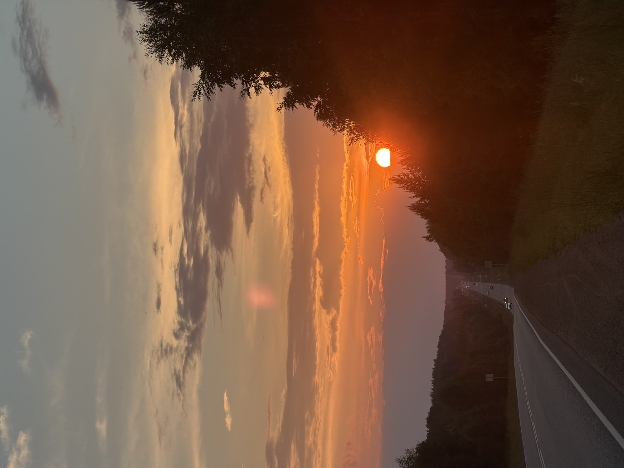 The image depicts a sunset over a highway in Chaikovsky, Russia. The sun, a bright orange orb, is low on the horizon, casting a warm glow across the sky. Wispy clouds are scattered across the sky, tinged with orange and pink hues. A dense line of trees borders the right side of the highway, silhouetted against the vibrant sky. The highway stretches into the distance, with a few cars visible, their headlights on as they travel along the road. The road surface is asphalt, with white lane markings. The surrounding landscape is hilly, covered in trees. The overall atmosphere is peaceful and serene, capturing the beauty of a rural sunset.