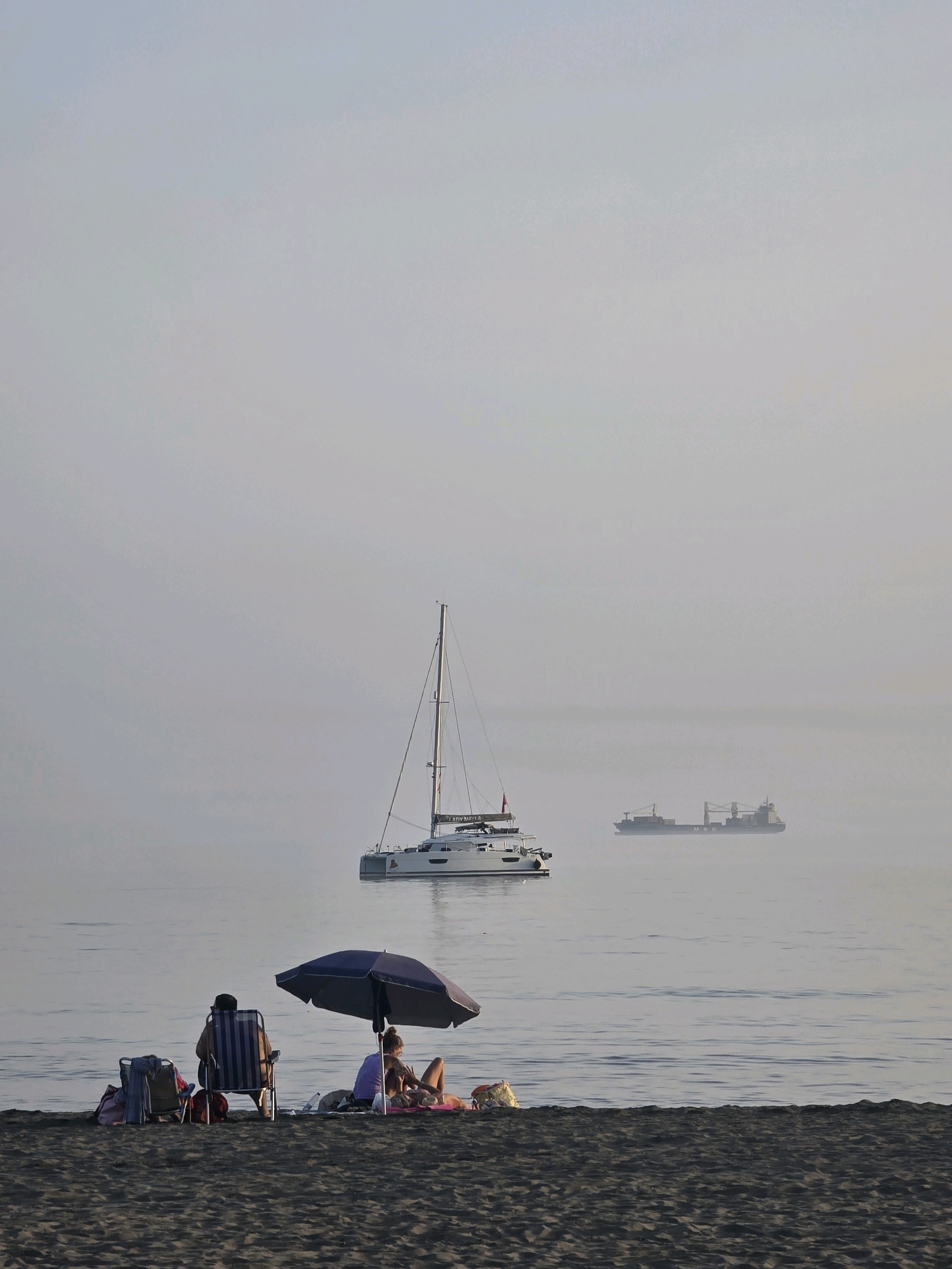 The image depicts a serene beach scene on a hazy day, likely in Málaga, Spain, given the presence of a sailboat and a cargo ship in the distance, common sights in the Mediterranean. In the foreground, on a dark sandy beach, two individuals are relaxing under a blue umbrella. One person is seated in a striped beach chair, facing away from the camera, while another person reclines on the sand, partially covered by a pink towel. Several beach bags and chairs are scattered nearby, suggesting a leisurely afternoon. In the middle ground, a white catamaran sailboat with tall masts is anchored on the calm, grey water. Further in the distance, a large cargo ship with the "MSC" logo is visible through the atmospheric haze, its outline softened by the mist. The overall atmosphere is tranquil, with the muted light and gentle waves creating a sense of peacefulness.
