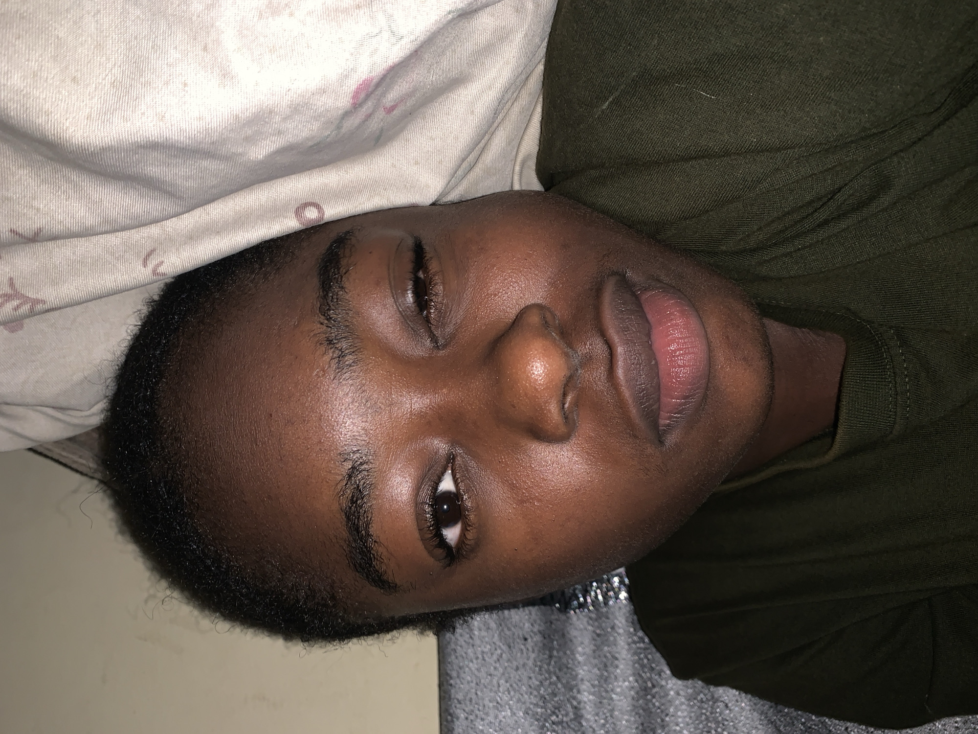 An individual with dark skin, short black hair, and dark eyebrows lies on their left side on a bed. The person wears an olive green t-shirt. The individual is positioned next to a white pillow with subtle designs. The individual's location in Rafin Par, Nigeria, is not determined from the image.