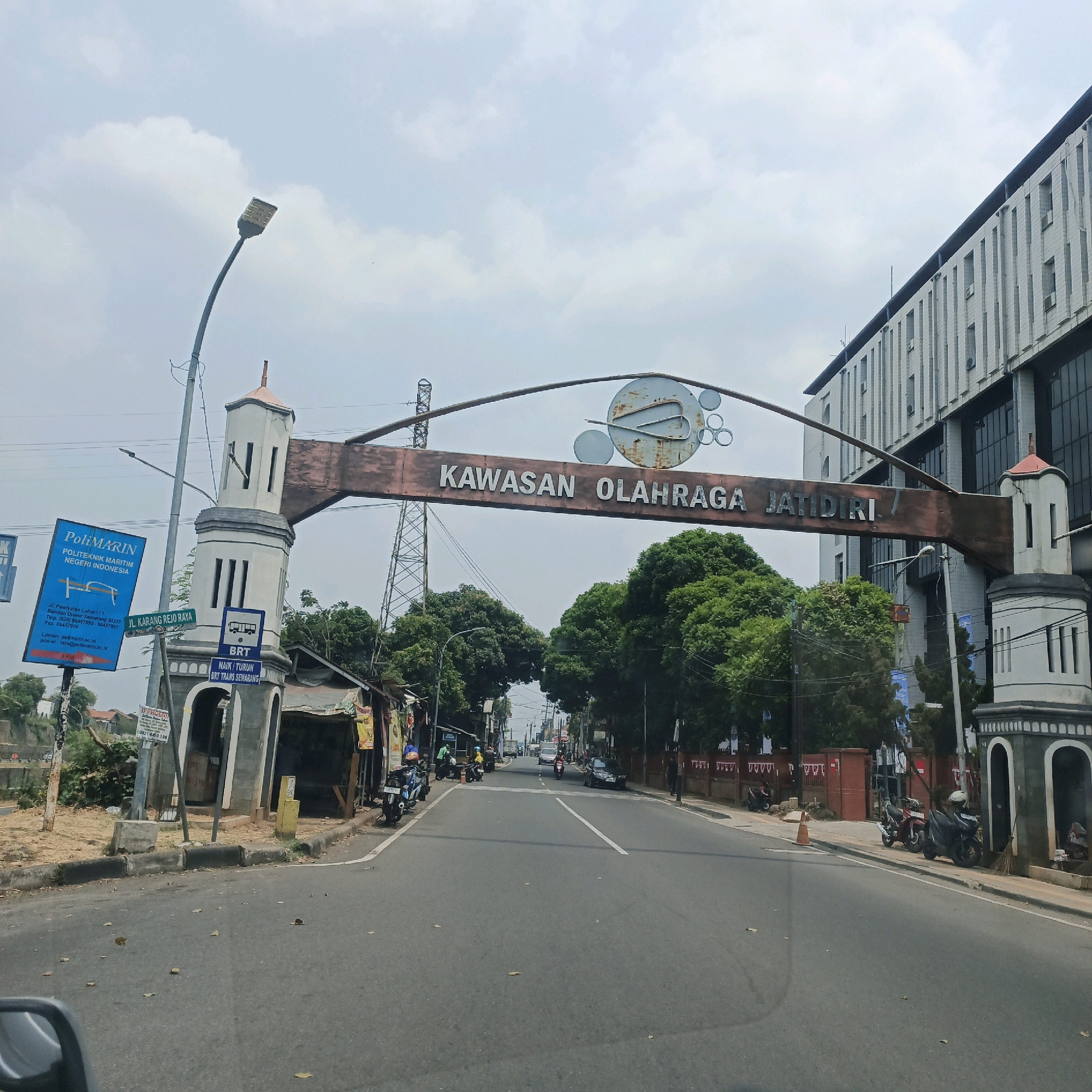 The image depicts a street scene in Semarang, Indonesia, likely at the entrance to the "Kawasan Olahraga Jatidiri" (Jatidiri Sports Area). A large archway spanning the road reads "KAWASAN OLAHRAGA JATIDIRI". On the left side of the road, there is a white tower structure with arches at the base, next to which is a blue sign for "POLIMARIN POLITEKNIK MARITIM NEGERI INDONESIA". Further down the road, a modern, multi-story building with a white facade is visible. The road is paved and has a white lane divider. Several motorcycles are parked along the side of the road. The sky is overcast with clouds. The scene appears to be during the daytime.