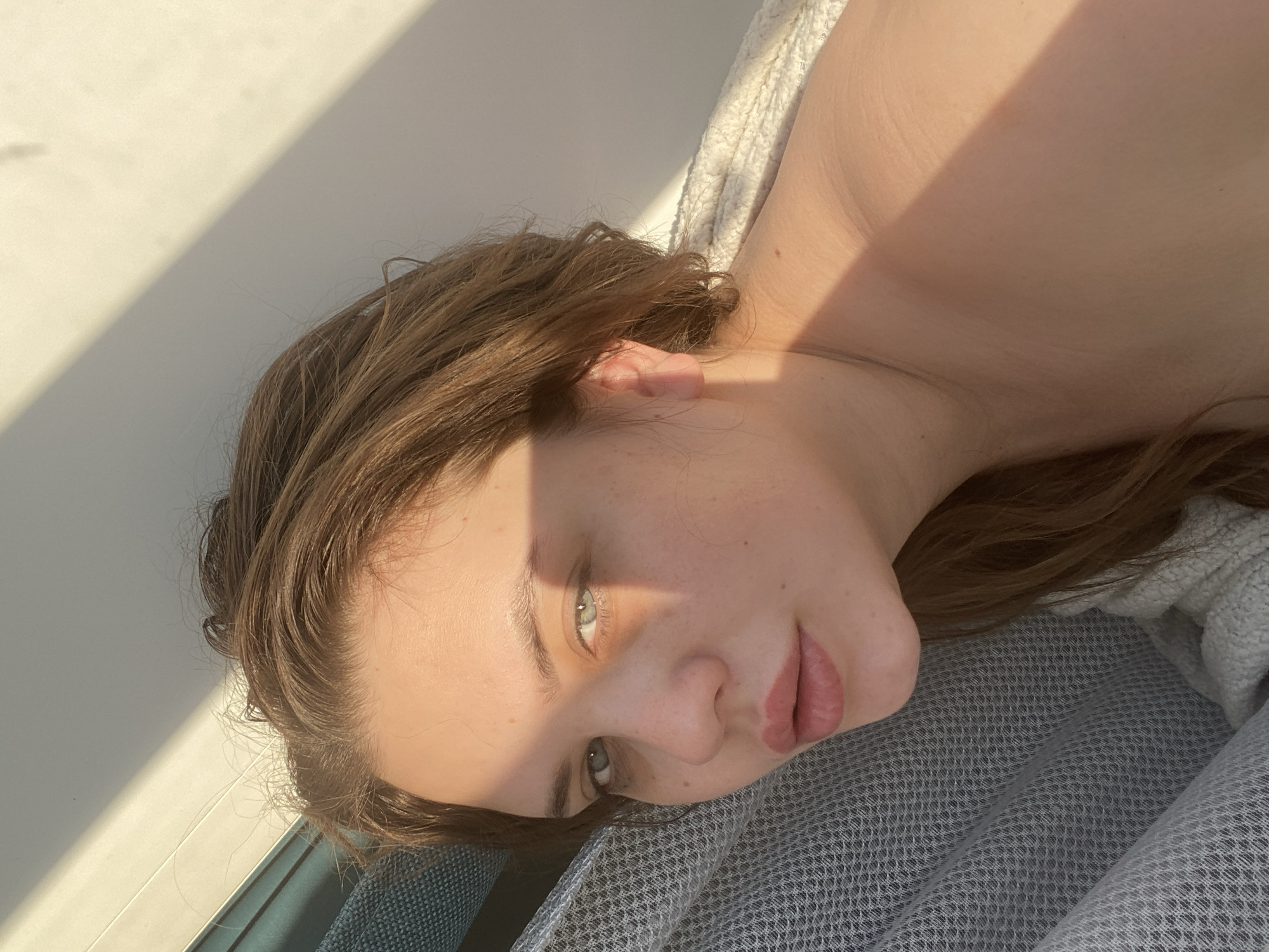 A woman with light brown hair and light eyes is lying down on a gray patterned surface. The photograph is taken at an angle, with her head tilted towards the bottom right corner. Part of her upper torso is visible, suggesting she may be topless. A white textured surface is seen behind her shoulder. A bright light source creates a sharp shadow across the left side of her face and onto the wall behind her. The scene's location in the city of Chernushka, Russia cannot be determined from this image.