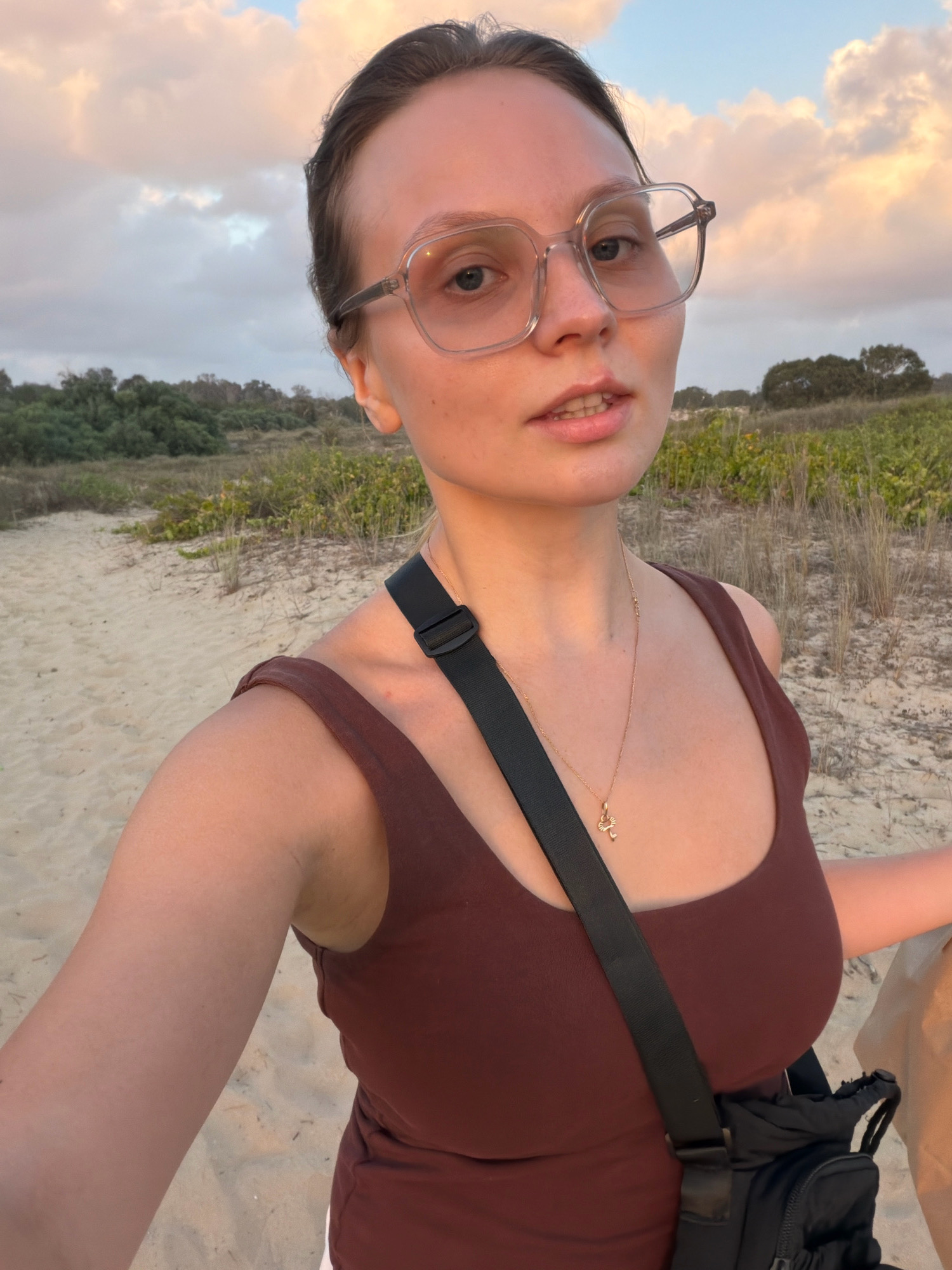 A young Caucasian woman with light skin and brown hair pulled back is taking a selfie outdoors. She is wearing large, clear-rimmed glasses, a dark brown tank top, and a gold necklace with a small pendant. A black strap from a bag is visible across her chest. She is holding a beige bag in her right hand. The background consists of a sandy path, green vegetation, and a cloudy sky with hints of orange and pink, suggesting it is either sunrise or sunset. There are no visible landmarks or indicators that place this image specifically in Rishon LeZion, Israel.