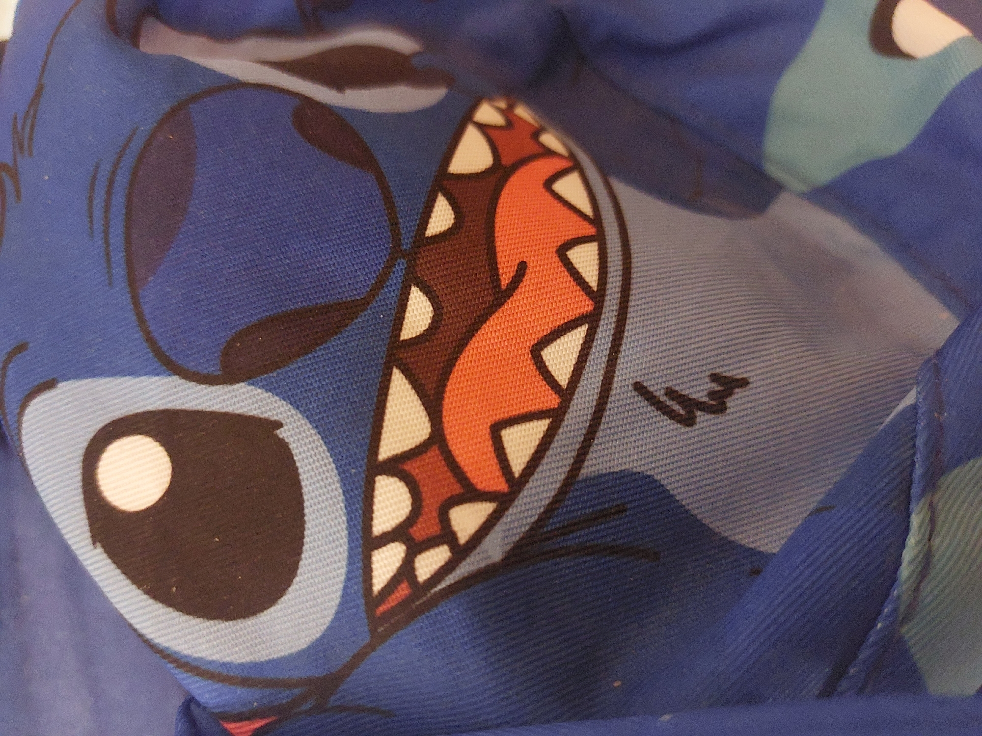 The image shows a close-up of fabric with a cartoon depiction of the character Stitch from the Disney movie "Lilo & Stitch". The fabric appears to be blue and includes details such as Stitch's eyes, mouth with sharp teeth, and some background elements of the design. There is no indication of the location being Tsarevo, Bulgaria, or any people, actions, or interactions in the provided image. The environment is limited to the fabric itself.