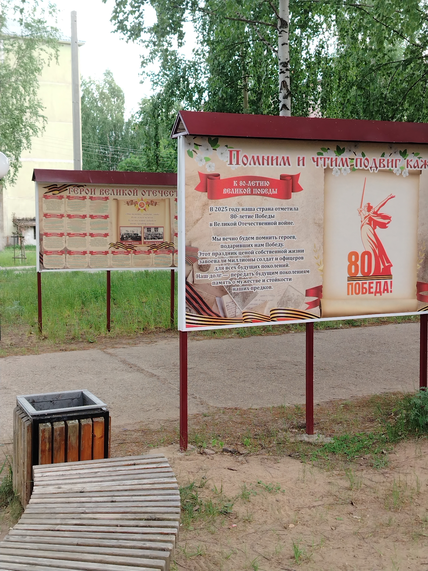 Here's a detailed description of the image:
**Setting:** The photo is taken outdoors in a park or similar public space, most likely in the city of Noviy, Russia, based on the language and imagery on the signs.
**Objects:**
* **Information Boards:** Two large information boards are visible. The right board commemorates the 80th anniversary of victory in the Great Patriotic War (World War II). It includes text in Russian and an image of the Motherland Calls statue. The left board has a headline: "Heroes of the Great Patriotic War".
* **Trash receptacle**: A square garbage can of wood and metal construction.
* **Bench**: A wooden bench is in the foreground.
**Text on the Boards:**
* Titles such as "80th Anniversary of the Great Victory".
* Sentences like, "In 2025, our country celebrated the 80th anniversary of Victory in the Great Patriotic War."
* Sentences celebrating the bravery of heroes.
* Sentences urging people to honor their ancestors.
**Environment:**
* A mix of pavement, dirt, and some grass.
* Trees in the background, including a birch tree.
* A multi-story building with visible windows.