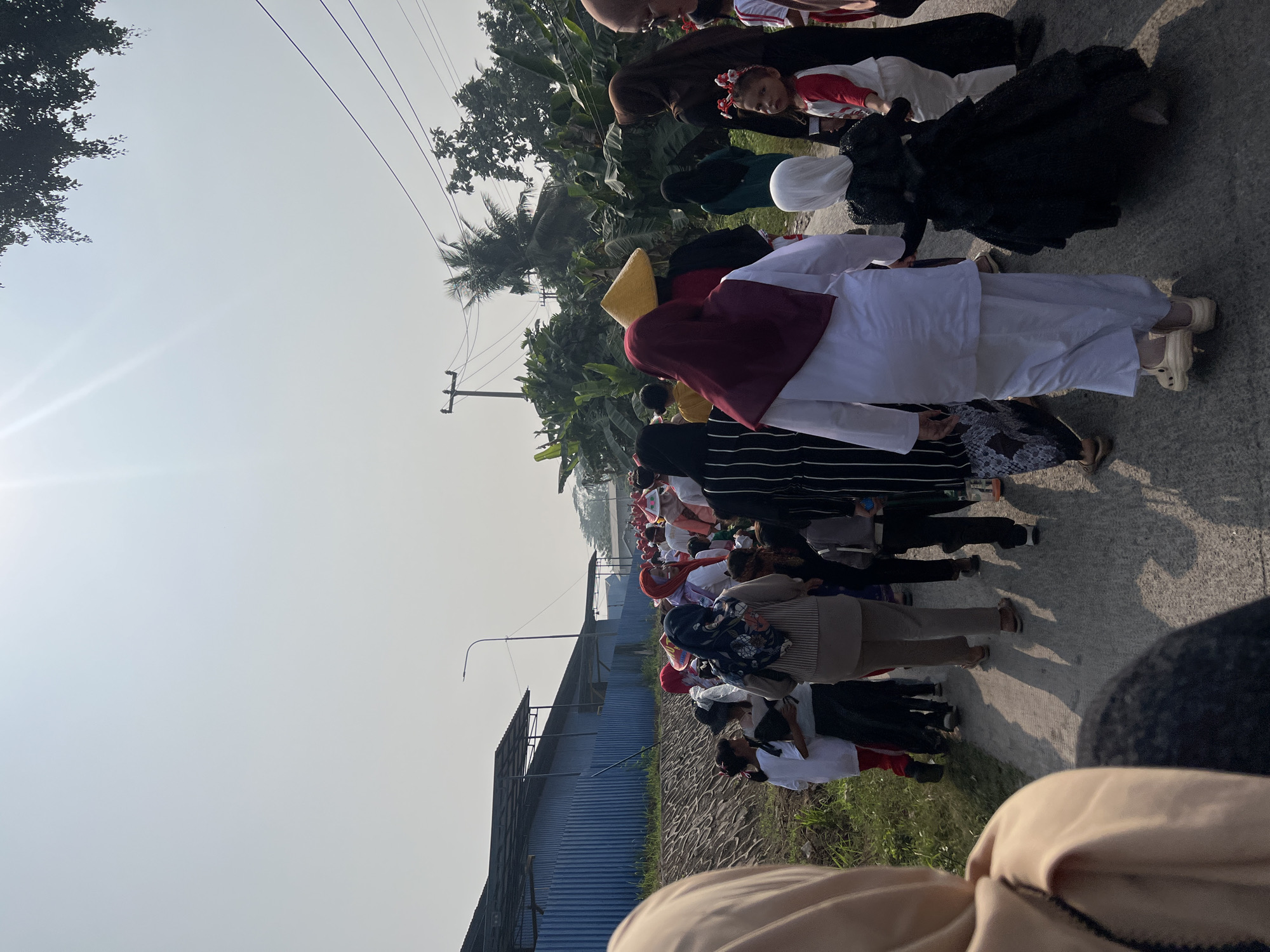 In Klapanunggal, Indonesia, a procession of people, predominantly women and children, walks along a paved road under a clear, hazy sky. The group appears to be participating in an event, with many wearing festive red and white hats, possibly indicating a Christmas or similar celebration, despite the tropical environment with abundant green foliage and palm trees lining the road. Some individuals are dressed in traditional Islamic attire, such as hijabs. A blue industrial building with a corrugated metal roof is visible on the left side of the road, bordered by a stone retaining wall and grass. In the background, utility poles and power lines are present, along with more vegetation and a few houses. The people are walking in the same direction, with some holding hands, suggesting a sense of community and shared activity. The lighting indicates it is daytime, with the sun casting long shadows on the road, implying it is either early morning or late afternoon.