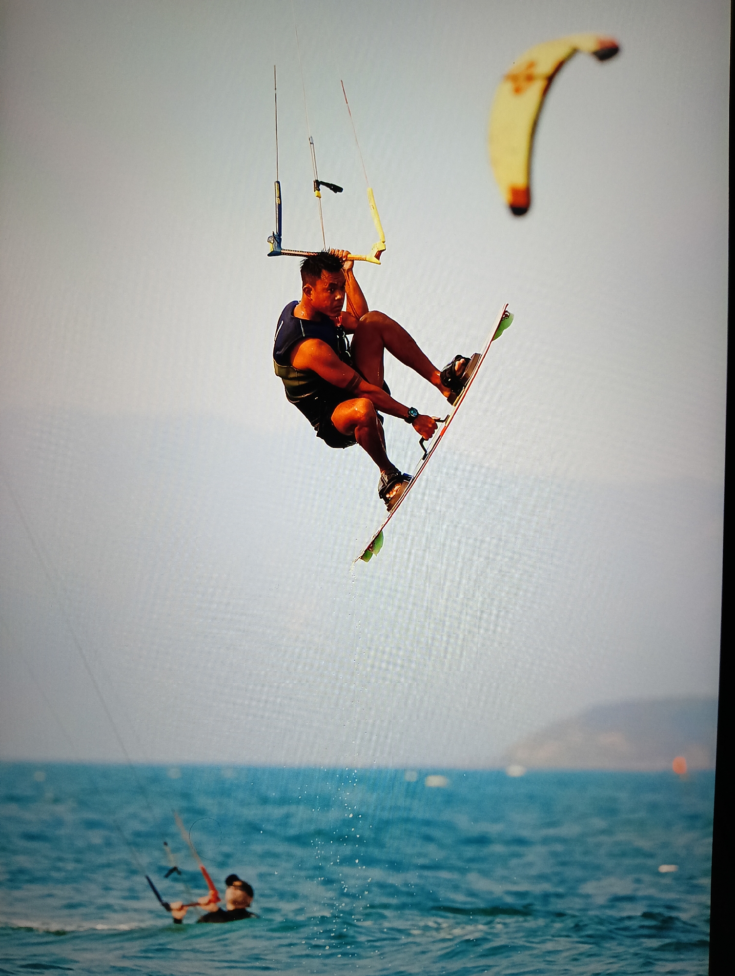 The image captures a man kitesurfing in the ocean. He is airborne, holding onto the kite's control bar with his right hand, and wearing a black vest and board shorts. His feet are strapped into a kitesurfing board. A yellow kite is visible in the background. Below the man, another individual in the water is holding a kite, with more of the ocean and distant landmass visible. The scene appears to be near Nha Trang Ward, Vietnam, given the terrain.