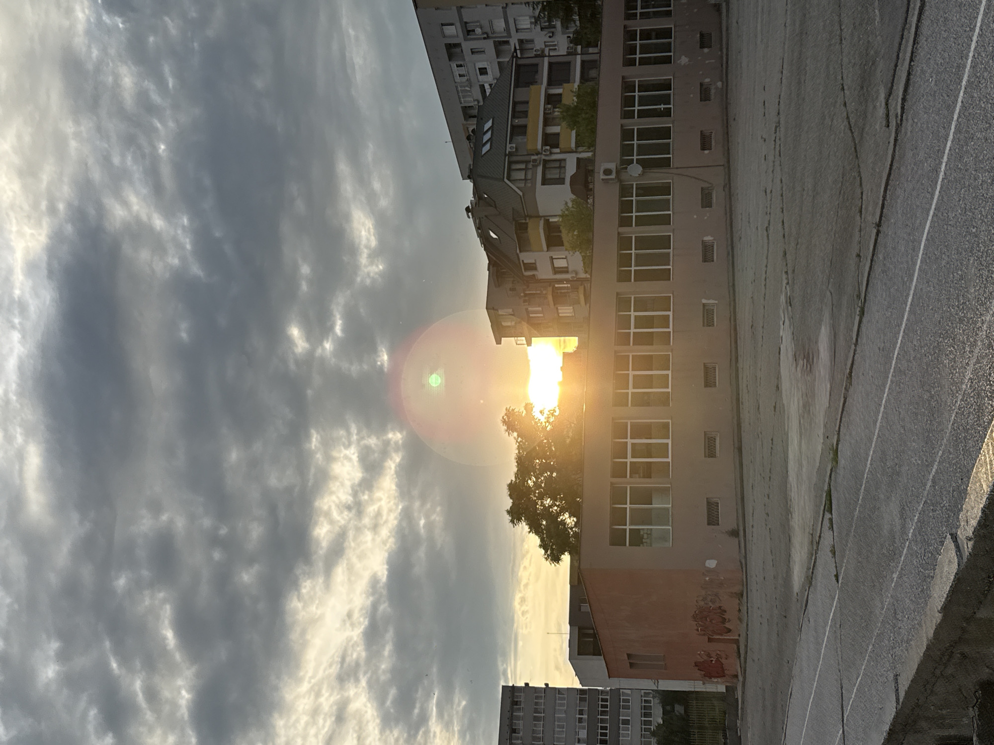 The image depicts an outdoor scene in Blagoevgrad, Bulgaria, likely during sunrise or sunset. In the foreground, a paved area with white lines, possibly a running track or sports court, is visible. Beyond this, a low, reddish-orange building with a long row of windows runs horizontally across the frame. Graffiti is visible on the left side of this building's wall.
Behind the low building, several apartment buildings of varying heights and architectural styles are visible. A prominent tree with green foliage stands in front of the buildings, partially obscuring the setting or rising sun, which casts a warm glow and creates a lens flare effect in the sky. The sky is filled with dramatic clouds, some dark and heavy, others lighter and illuminated by the sun. The overall impression is a somewhat urban landscape bathed in the atmospheric light of dawn or dusk. There are no people or animals visible in the image.