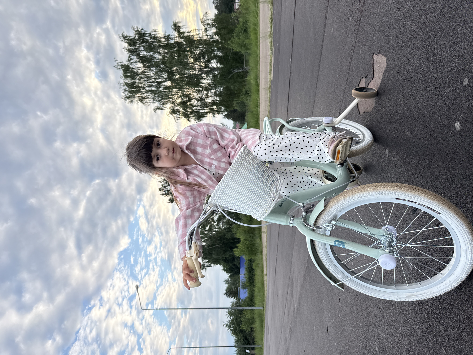 An outdoor medium shot captures a young girl seated on a light green bicycle with training wheels. She is positioned in the center of the frame, looking directly at the camera with a neutral expression. The girl wears a pink and white checkered long-sleeved shirt and white pants with black polka dots. Her right hand rests on the bicycle's handlebars. The bicycle features a white woven basket on the front and white tires. The background consists of a paved surface, a line of trees, and a sky with scattered clouds. Two streetlights are visible in the distance to the left. The image appears to have been taken in Chaikovsky, Russia, given the presence of birch trees common to the region and the general aesthetic of the environment.