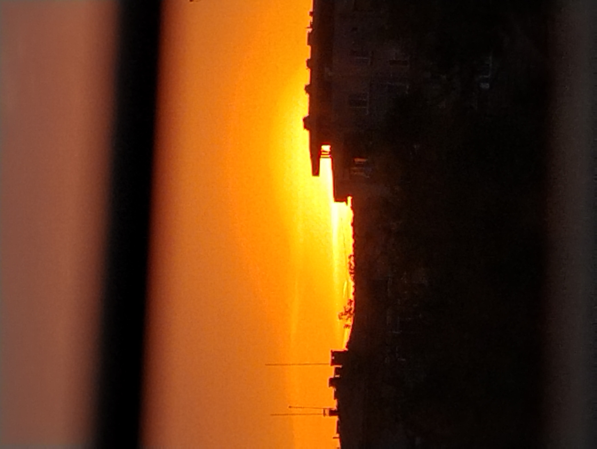 The image depicts a sunset in an urban area. The sky is a gradient of orange and yellow. In the foreground, a building and several radio towers are silhouetted against the sky. Vertical elements frame the left and right edges of the scene. The city and state are Unknown.